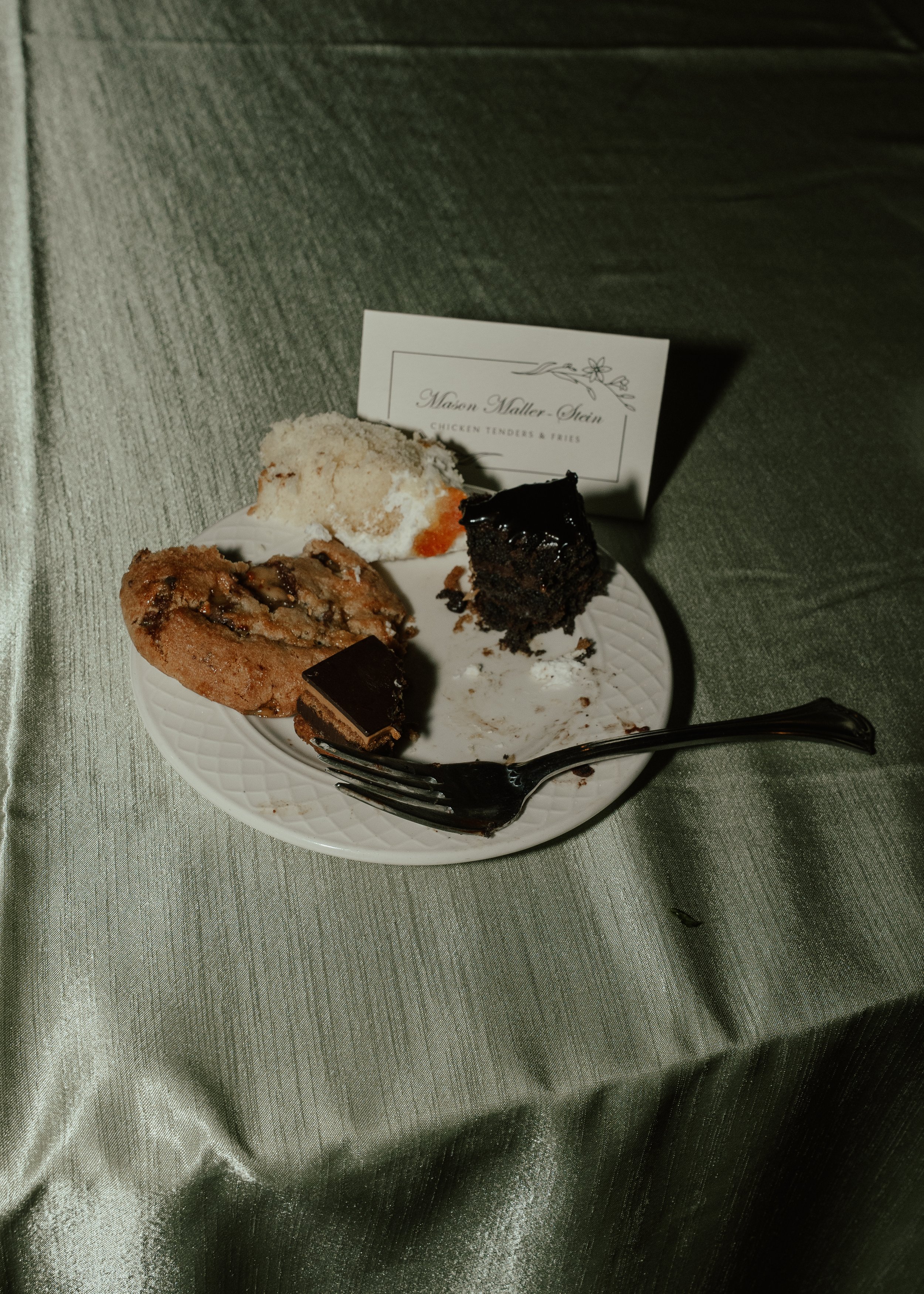 A white plate with three pieces of dessert, a fork, and a card nearby. One dessert is a bite-sized chocolate cake with glaze, another is a cookie with chunks, and the third is a slice of cake with white frosting. The card reads 'Mason Maller-Stern' a