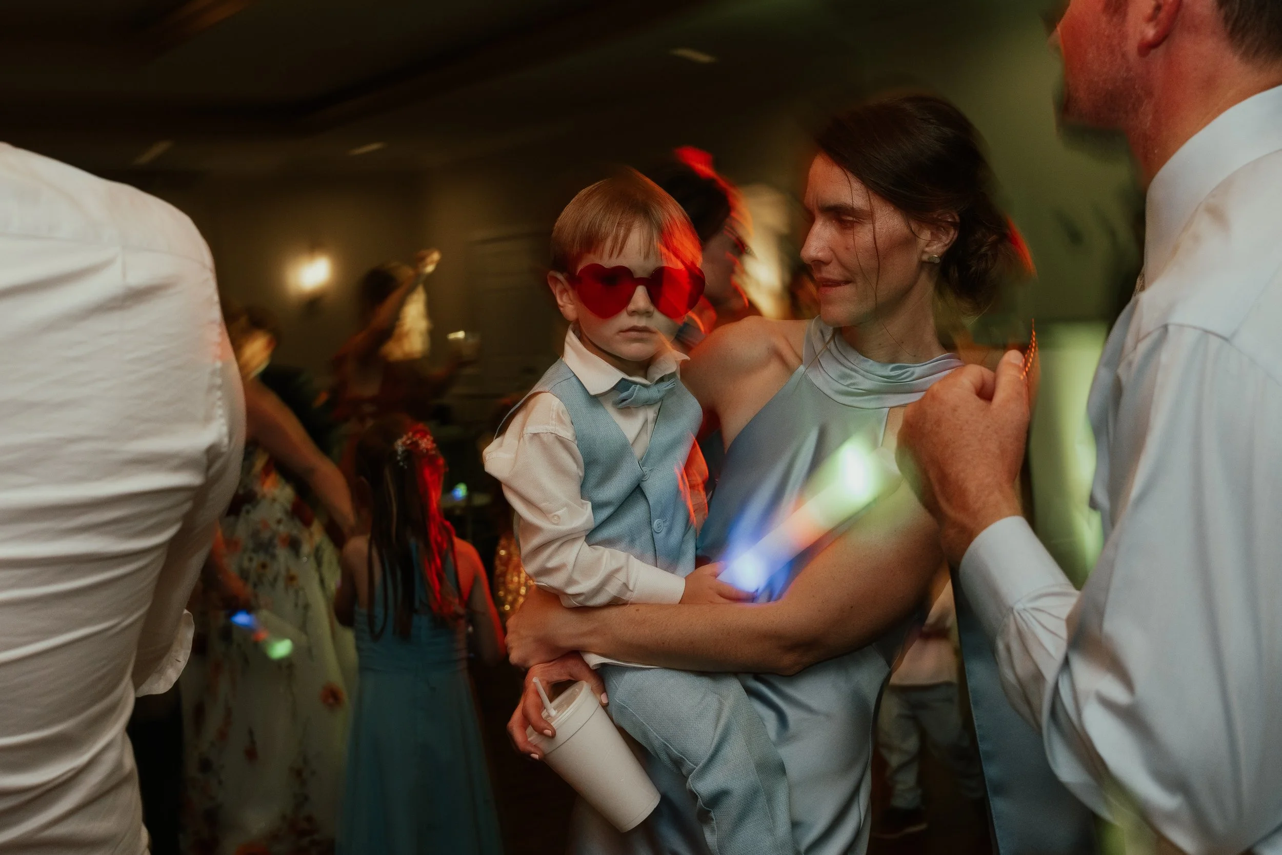 A woman dancing with a young boy wearing red heart-shaped sunglasses at a party or celebration with other people dancing in the background.
