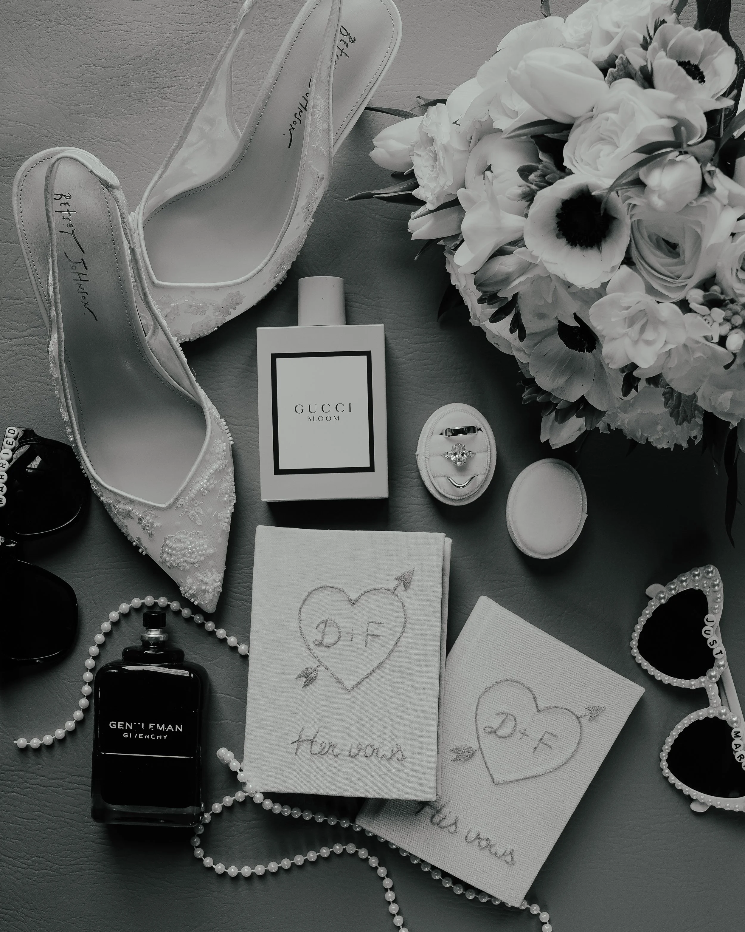 A collection of wedding items on a table, including white high-heeled shoes, a bouquet of white flowers, perfume bottles, a pearl necklace, sunglasses, and paper napkins with vows written on them.