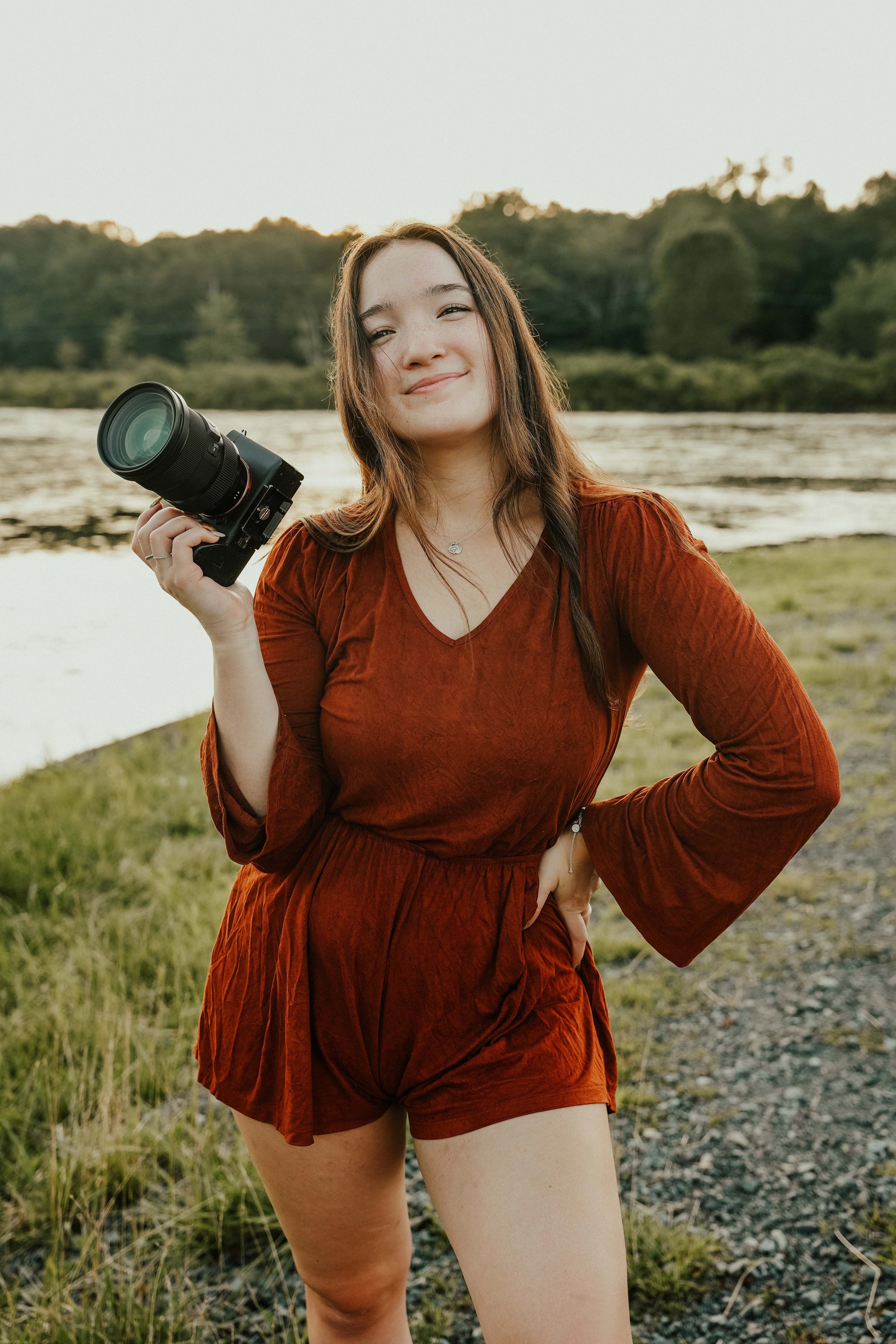 A young woman outdoors near a river at sunset, holding a camera and smiling, with her hand on her hip, wearing a rust-colored outfit.