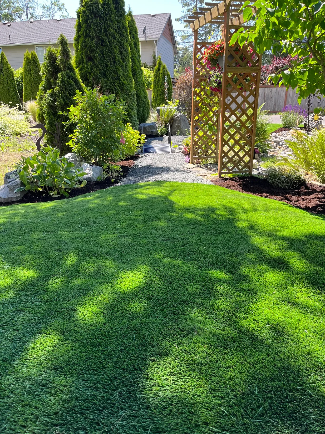 A backyard garden with a lush green lawn, surrounded by trees, shrubs, and flowering plants, with a wooden trellis on the right and a stone pathway leading to a small bridge in the background.