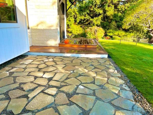 Stone patio with cracked pattern in a backyard, surrounded by green grass, trees, and garden furniture along a fenced yard.