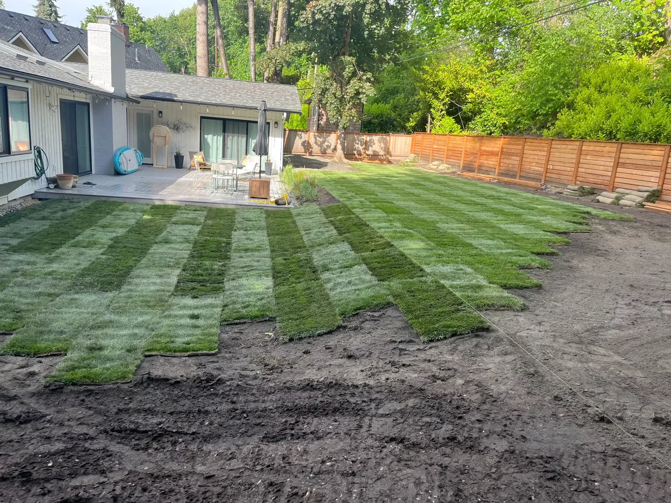 A backyard with freshly laid sod grass in a checkered pattern, an outdoor patio with furniture and a closed umbrella, a wooden fence, and trees providing shade.