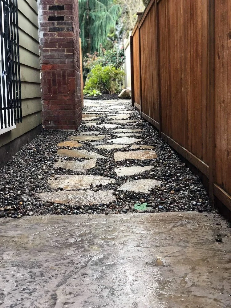 A narrow outdoor pathway with flagstone stepping stones and gravel, bordered by a house with horizontal siding on the left and a wooden fence on the right, leading into a lush garden area.