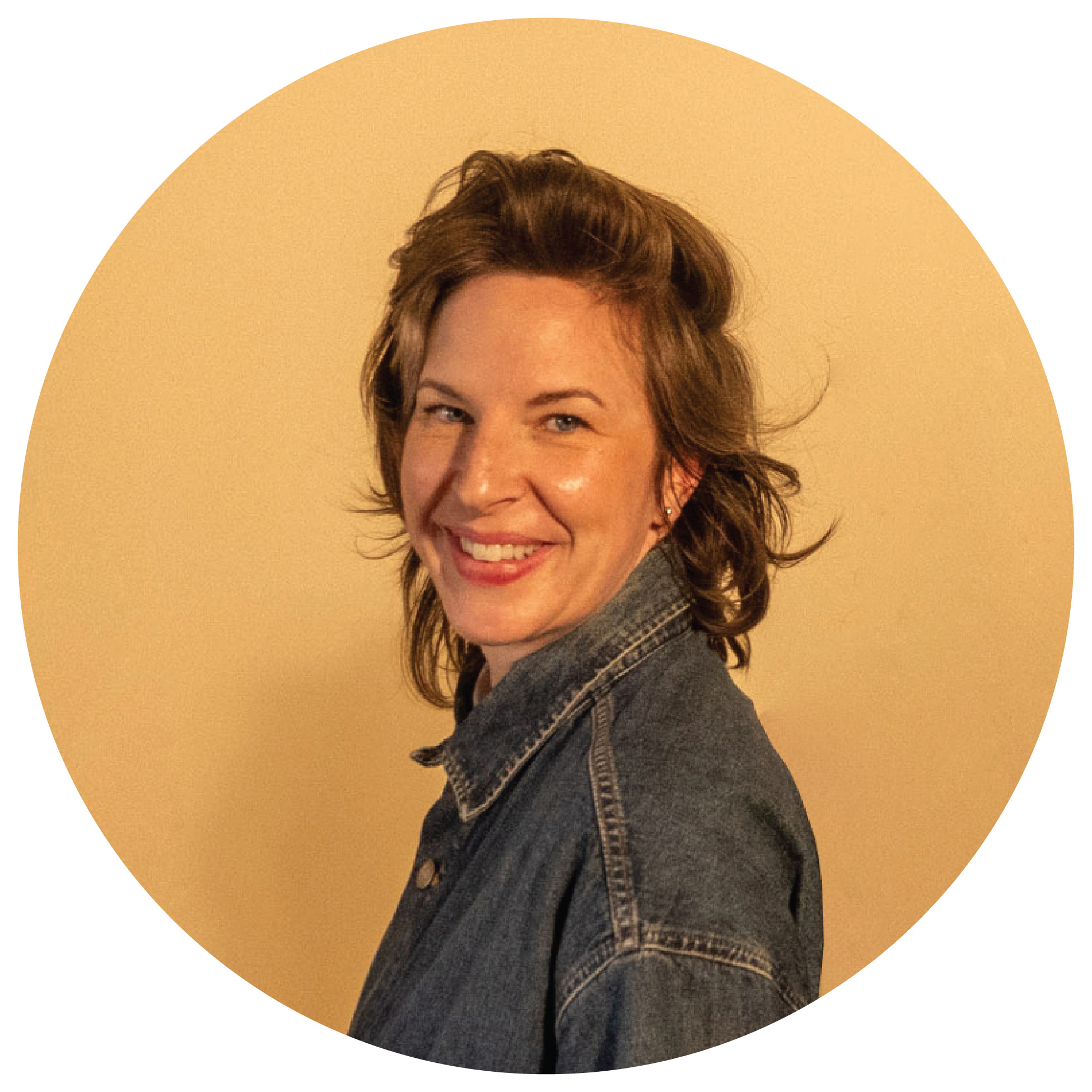 A woman with brown hair smiling and wearing a denim jacket, facing sideways against a beige wall.