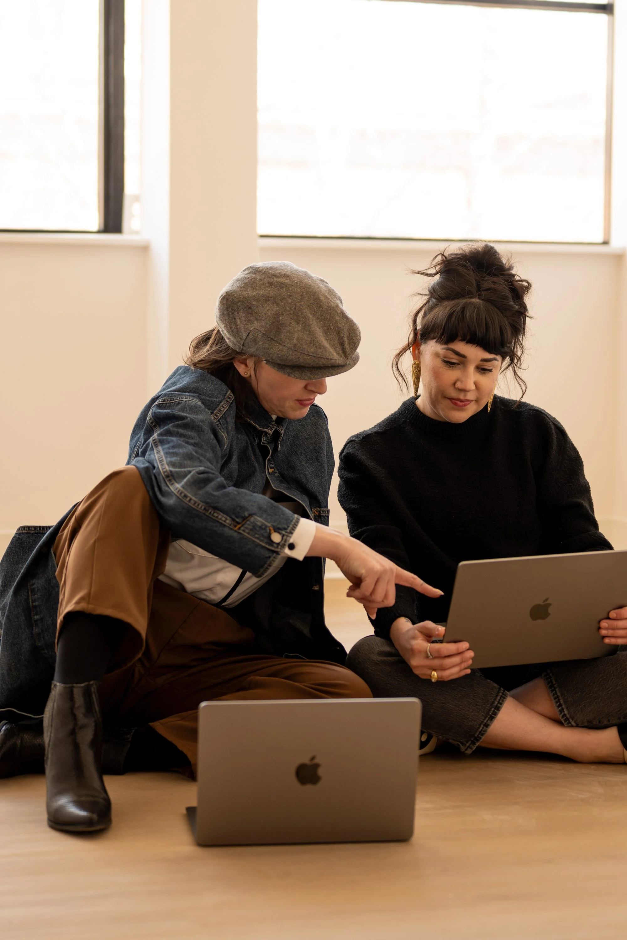 Two women sitting on the floor with laptops, one is pointing at the laptop screen the other is looking at it.