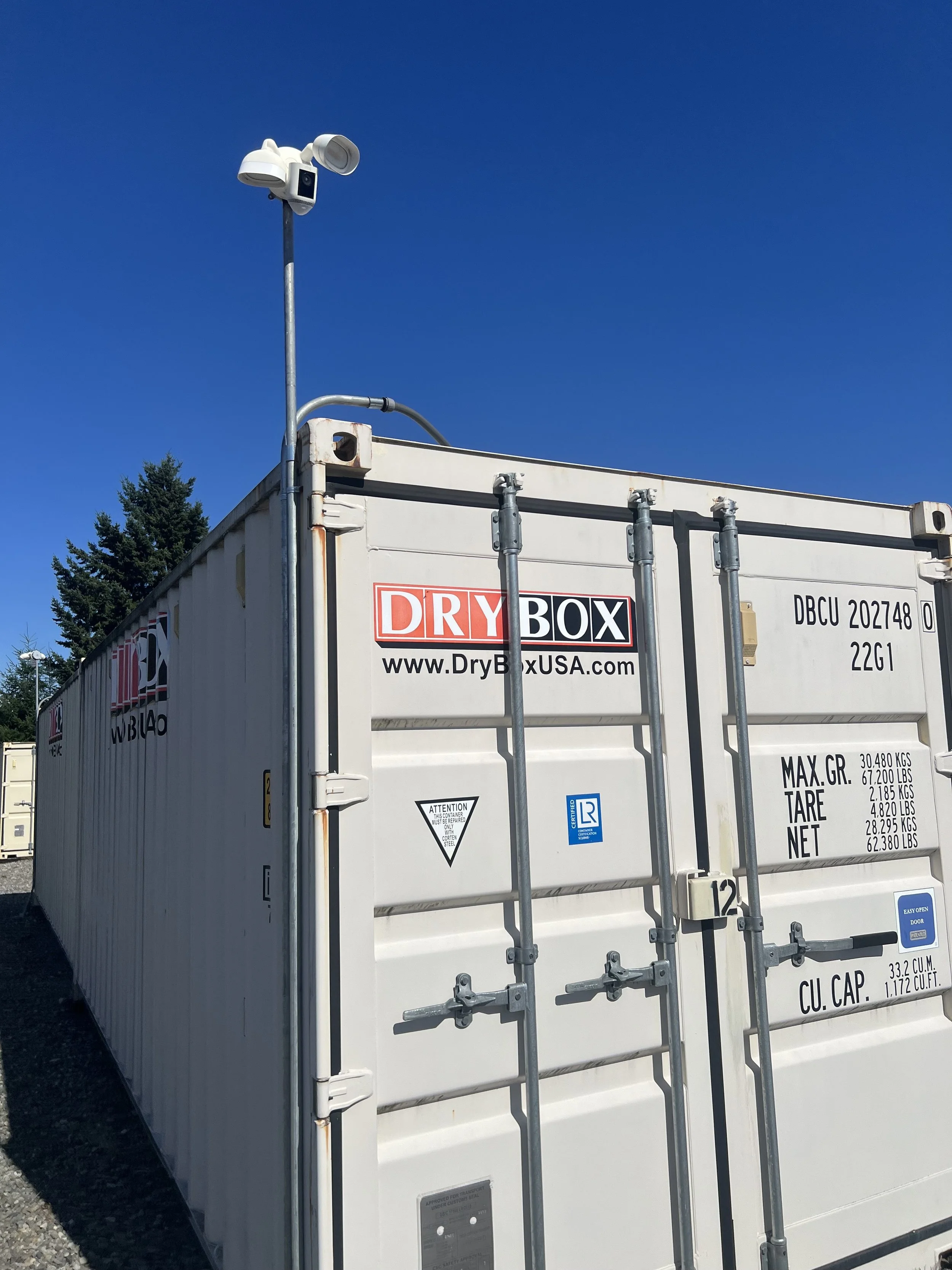 A white dry storage container with a security camera mounted on a pole above it, set against a clear blue sky. The container has signs and labels including a logo for DryBoxUSA.