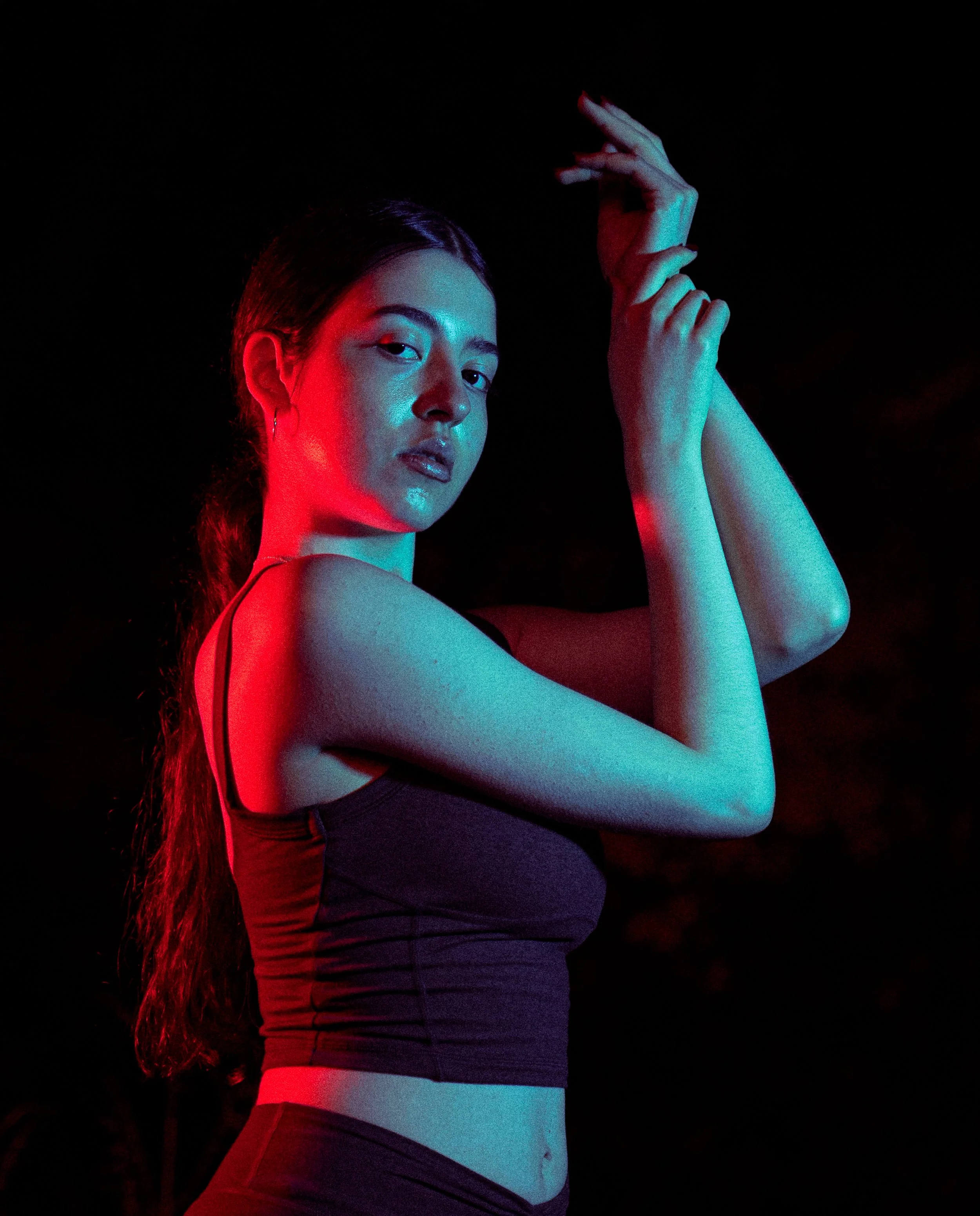 Young woman with long hair styled in a ponytail in low light, illuminated with red and blue lighting, posing with arms raised and crossed in front of a dark background.