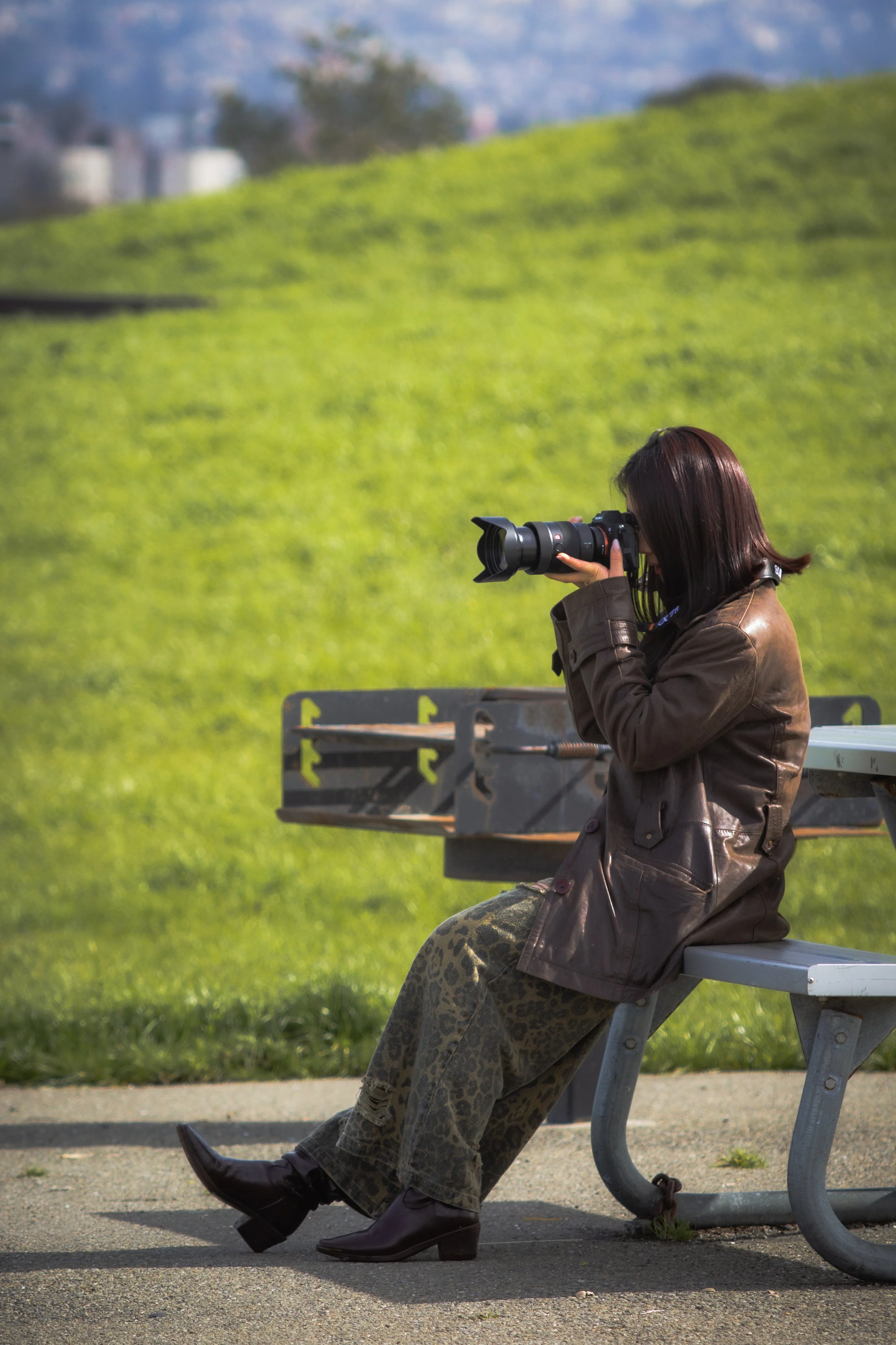 Woman in brown leather jacket and leopard print pants sitting on park bench, taking photograph with a camera.
