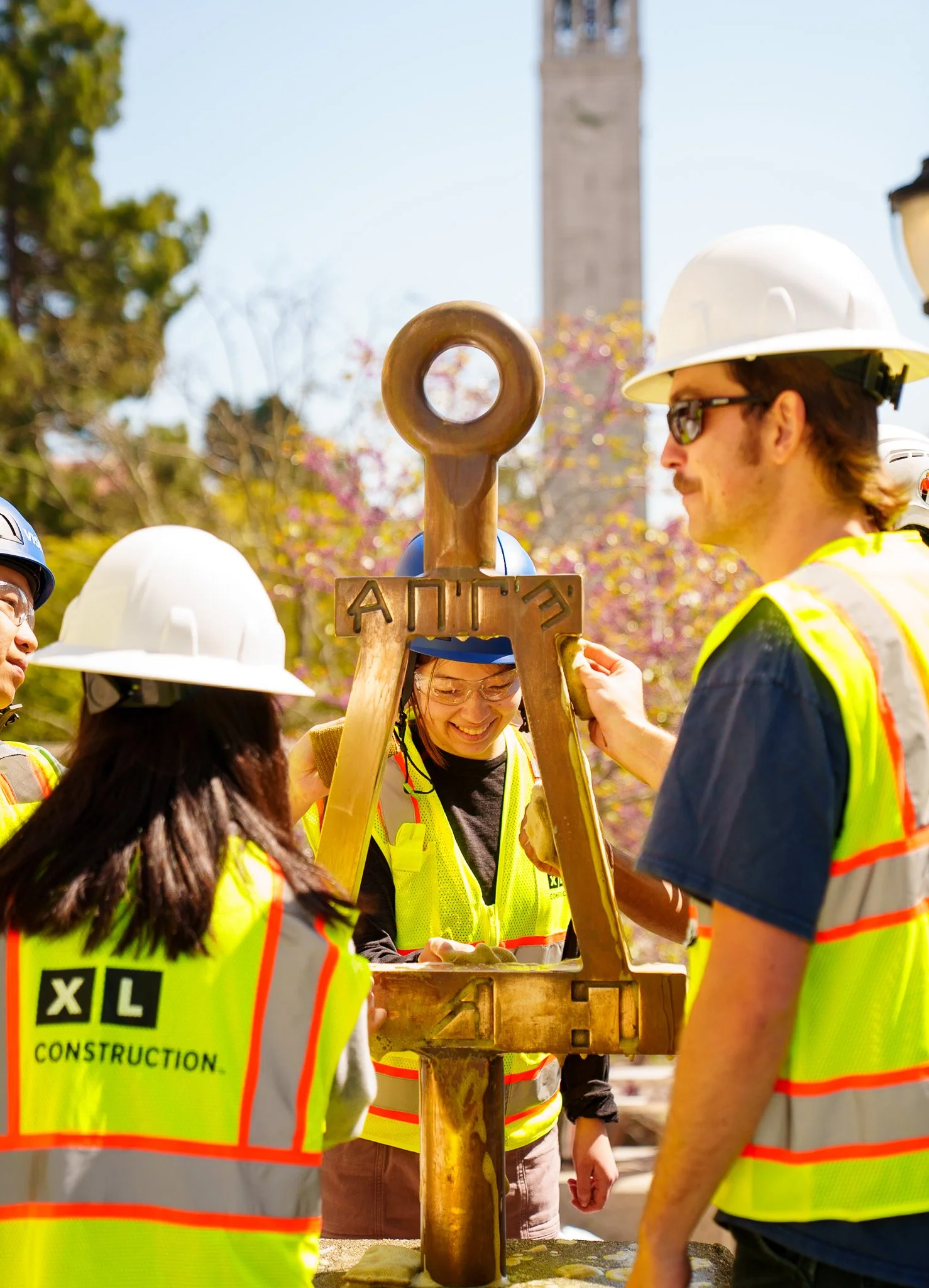 Construction workers in safety vests and helmets smiling and working with a large bronze anchor at an outdoor construction site.