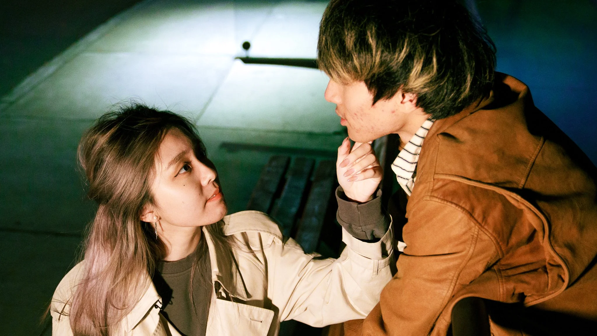 A young woman and young man are facing each other closely indoors, with the young woman sitting and the young man leaning over her. The woman has light brown hair and is looking up at the man with a serious expression. The man has dark hair and is resting his chin on his hand, looking down at her.