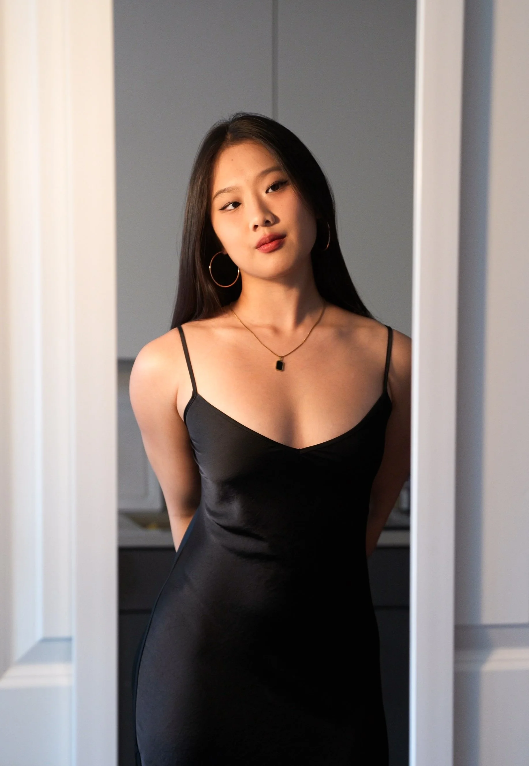 A young woman with long black hair wearing a black spaghetti strap dress, standing in a doorway and looking at the camera.