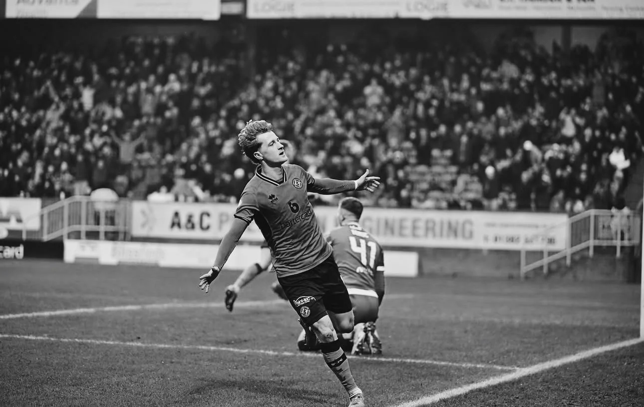 A soccer player celebrating a goal on the field, with a crowd in the stands behind him.
