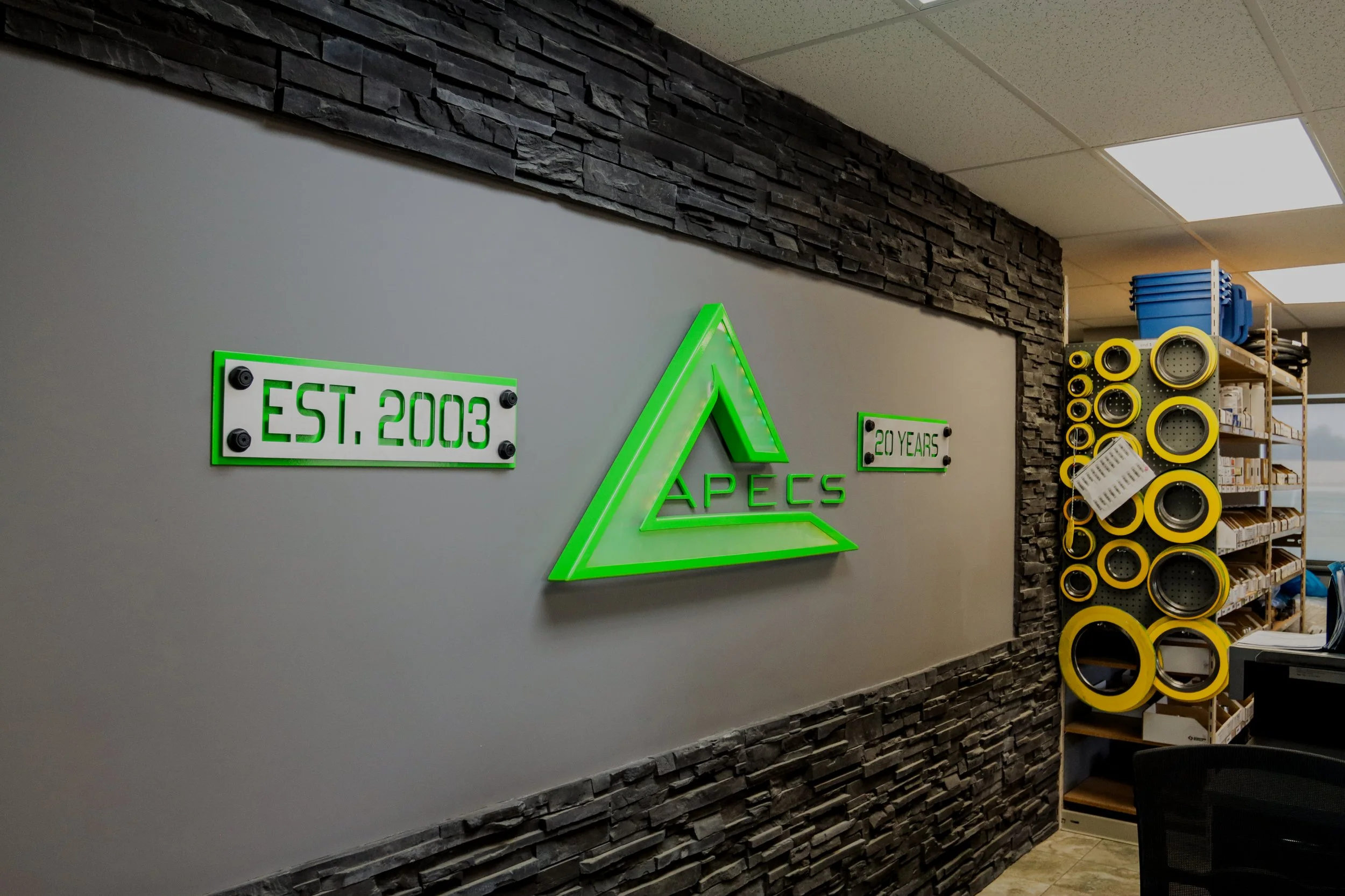 Interior office wall with illuminated green and white sign that reads 'EST. 2003' and '20 YEARS' flanking a stylized green triangle with the word 'APECS' inside. To the right, a shelf with yellow and black speakers and other hardware in a workspace.