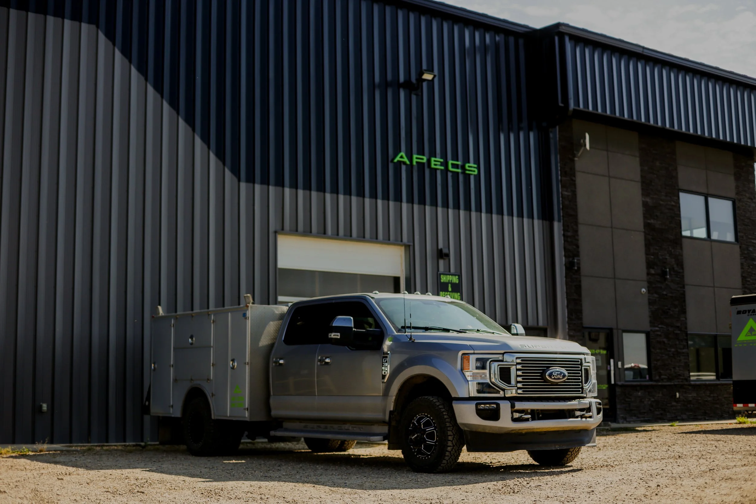 A silver Ford pickup truck parked outside a commercial building with dark gray siding and large windows. The building has a green sign that reads 'APECS' and another smaller sign near the door that says 'Shipping & Receiving.'
