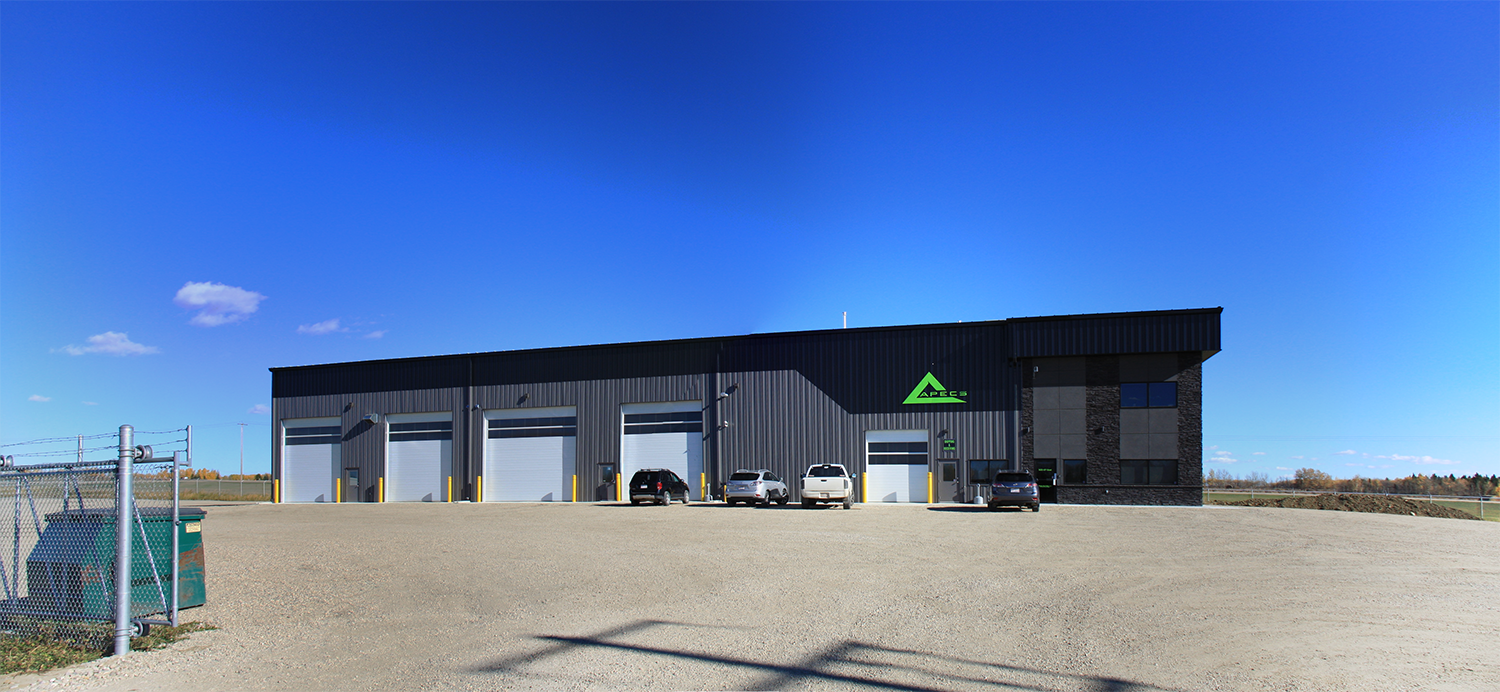 Industrial warehouse building with five large garage doors, parked cars in front, and a sign that reads 'APECS' under a blue sky.