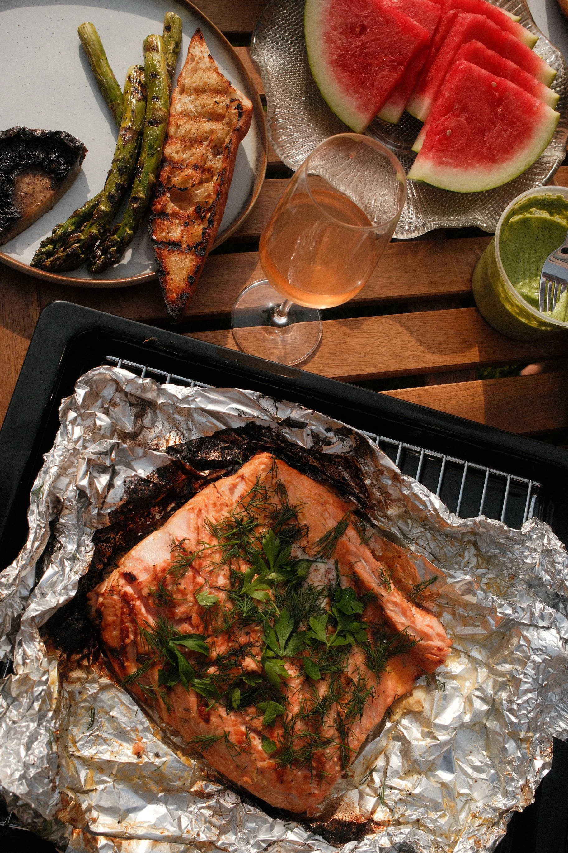 Grilled salmon with herbs on baking sheet, accompanied by grilled asparagus and salmon steak on a plate, sliced watermelon on a glass plate, a glass of rosé wine, and a green sauce in a jar.