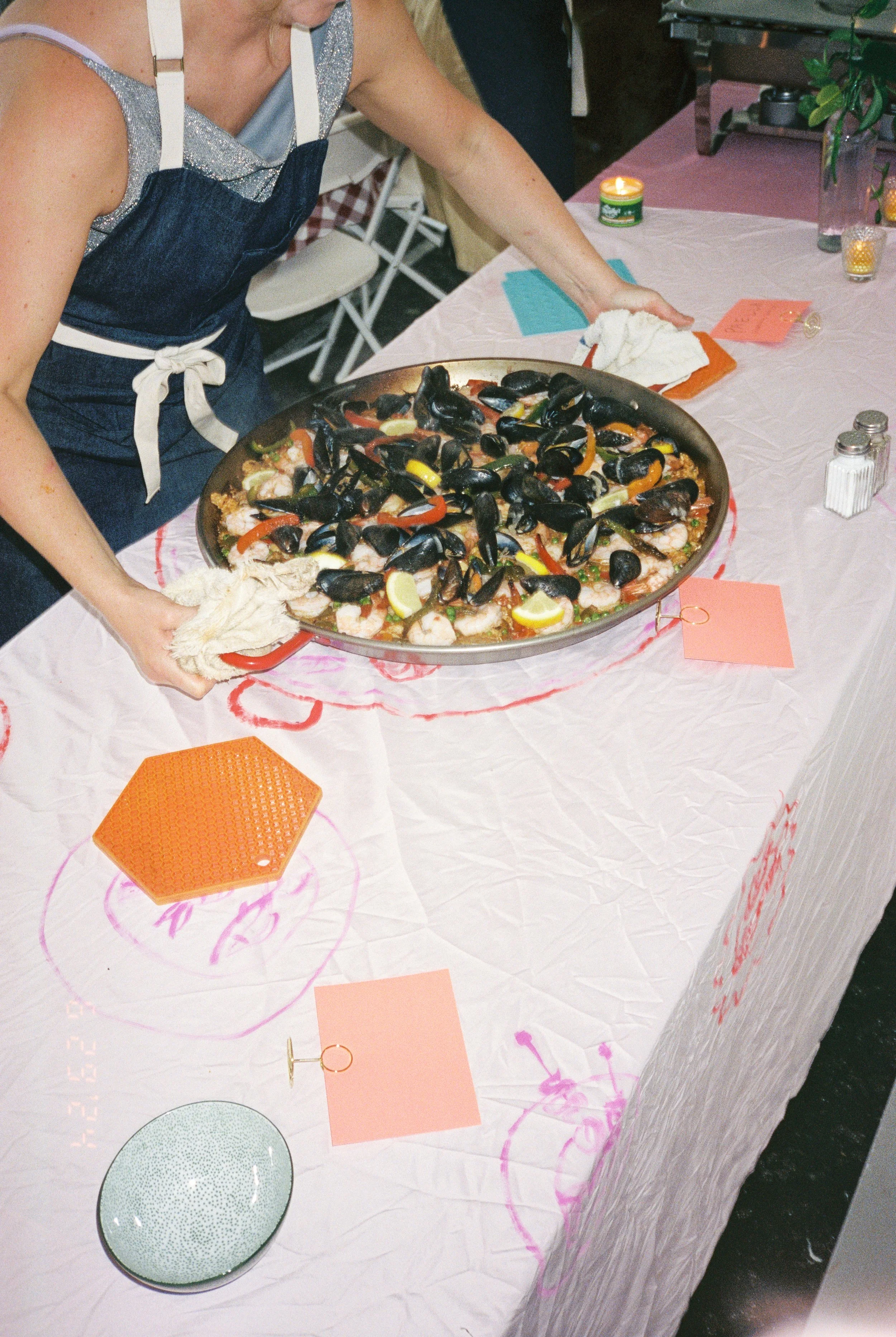 A person wearing a blue apron and a silver top arranging a large tray of cooked mussels, shrimp, and lemon slices on a table with pink and orange decorations, salt and pepper shakers, and a lit candle.