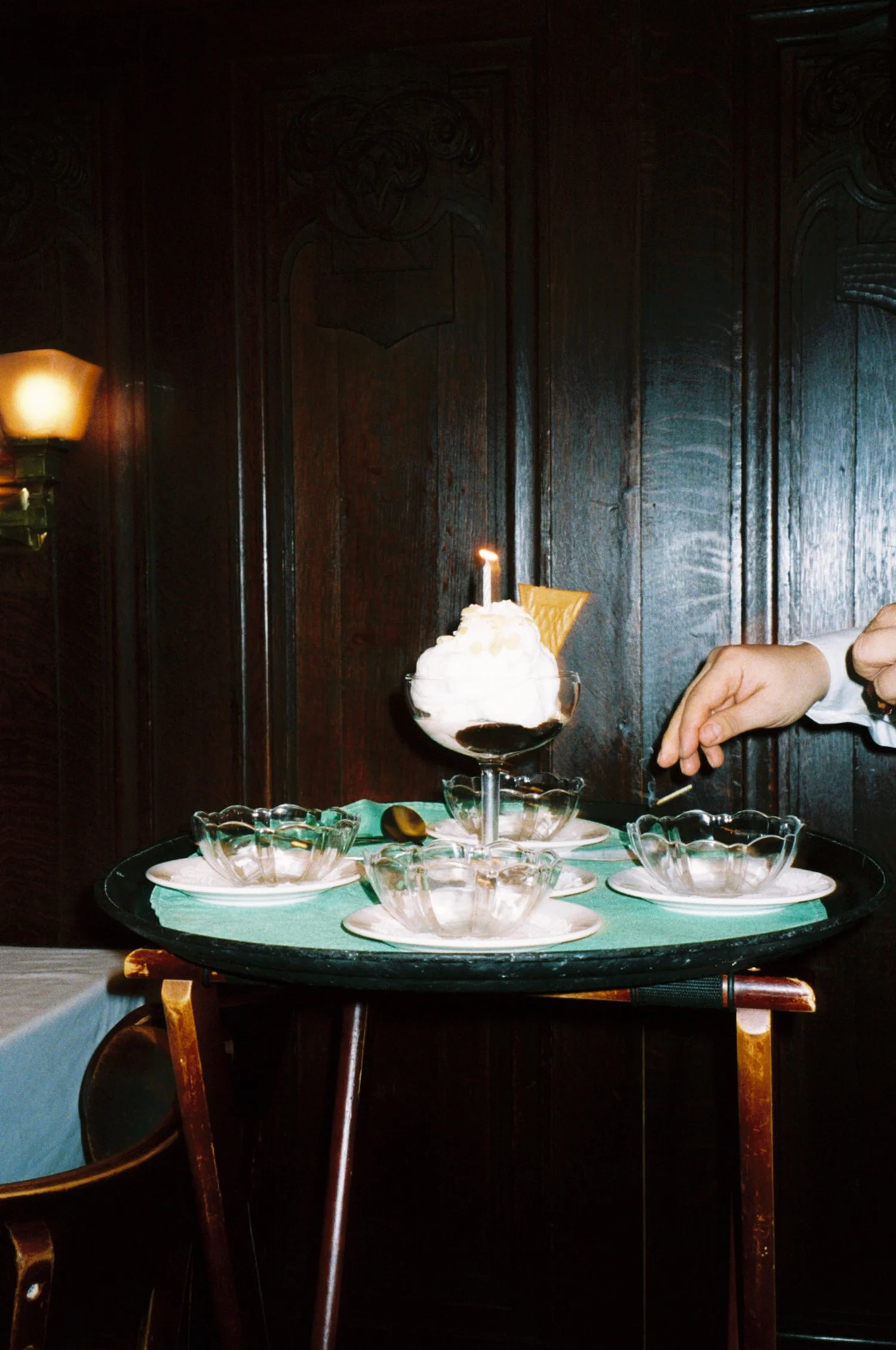 A glass dessert cup with whipped cream, a lit birthday candle, and a triangular wafer, surrounded by glass bowls, on a black tray with a green cloth, set on a wooden table with dark wood-paneled walls in the background.
