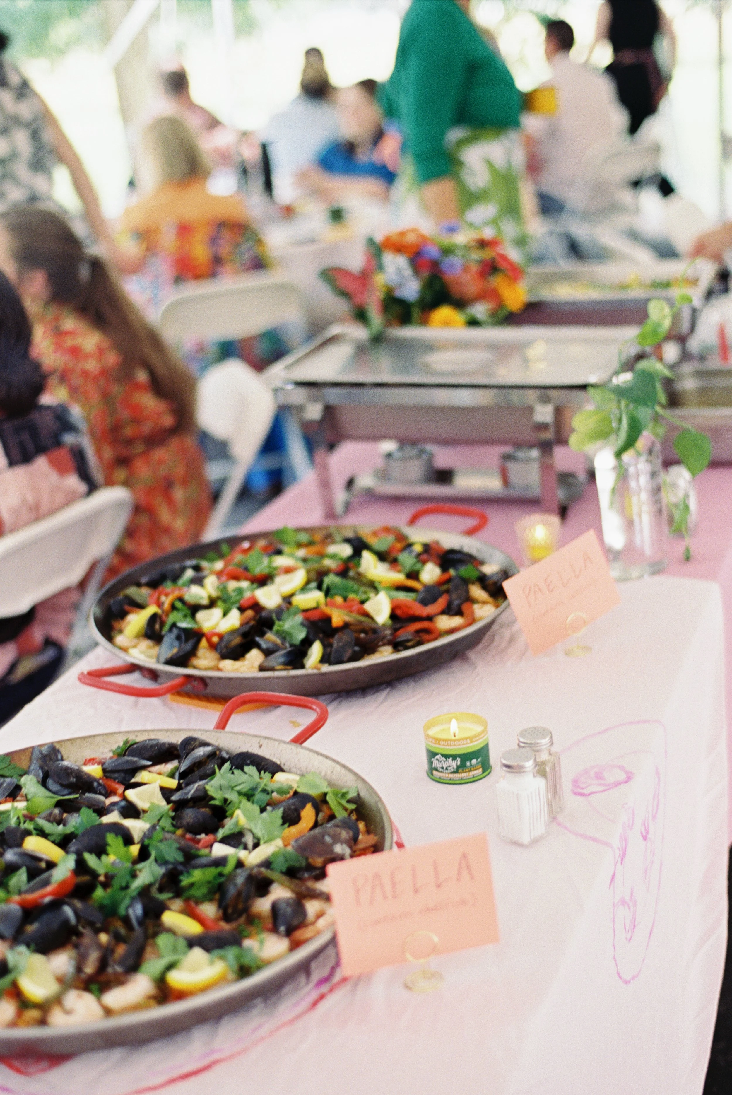 Two large pans of colorful seafood paella on a table at a gathering, with a pink sign labeled 'PAELLA' and people in the background.