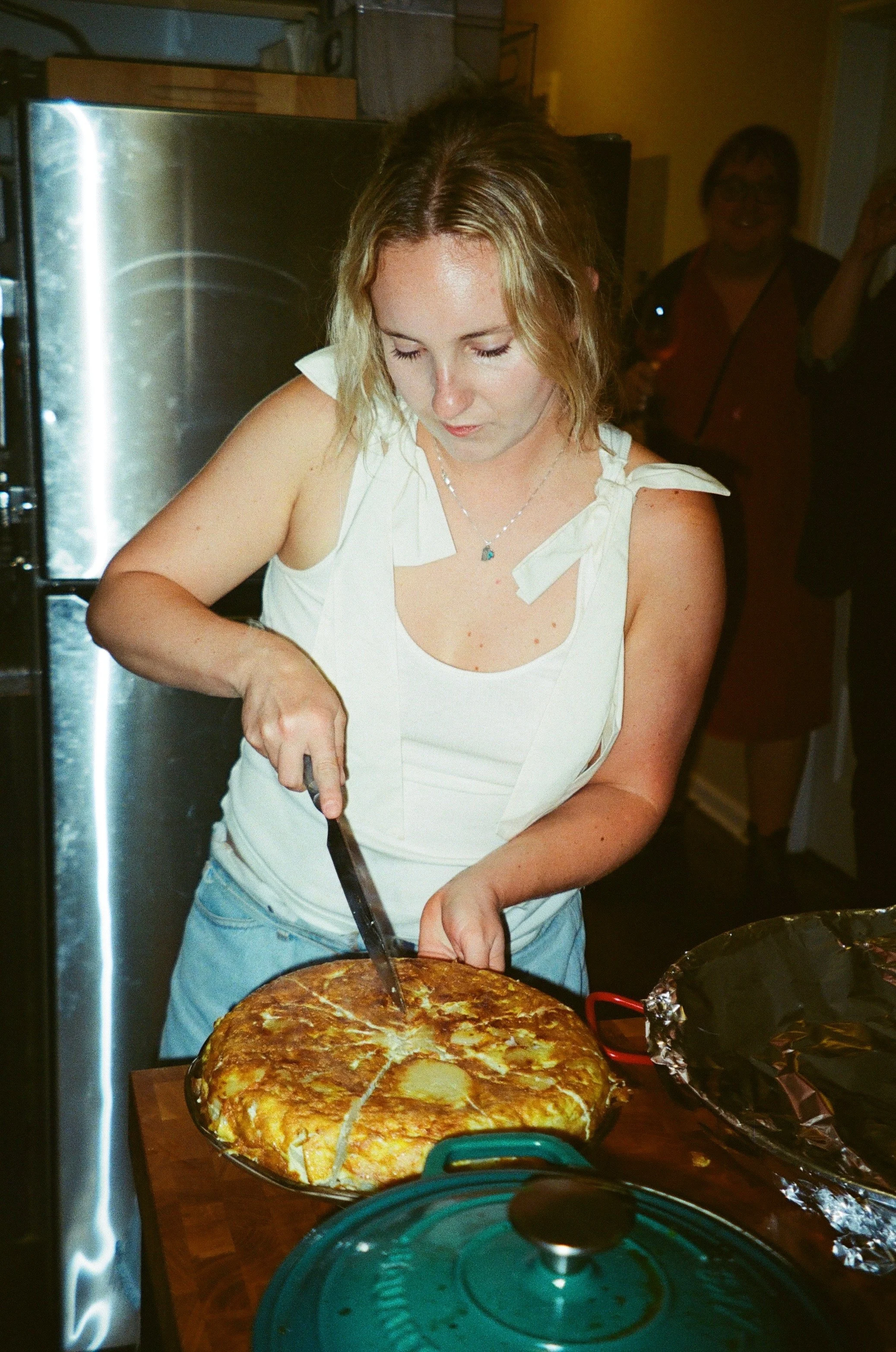 A woman with blonde hair slicing a large pizza with a knife in a kitchen.