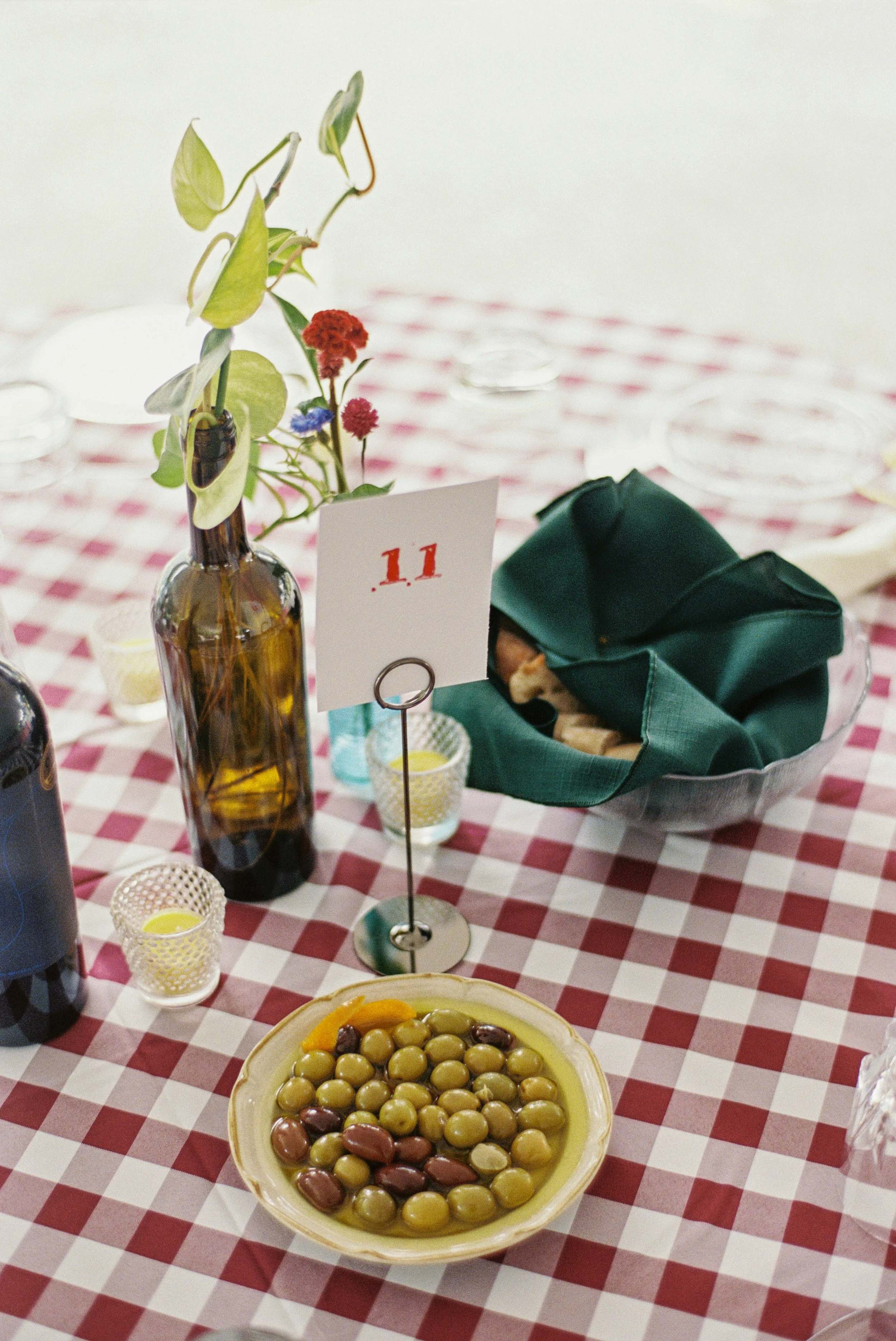A table with a red and white checkered tablecloth features a ceramic bowl filled with green and black olives, a small bowl with yellow sauce, a tall amber glass bottle with mixed flowers, small candle holders with lit candles, a card with the number 