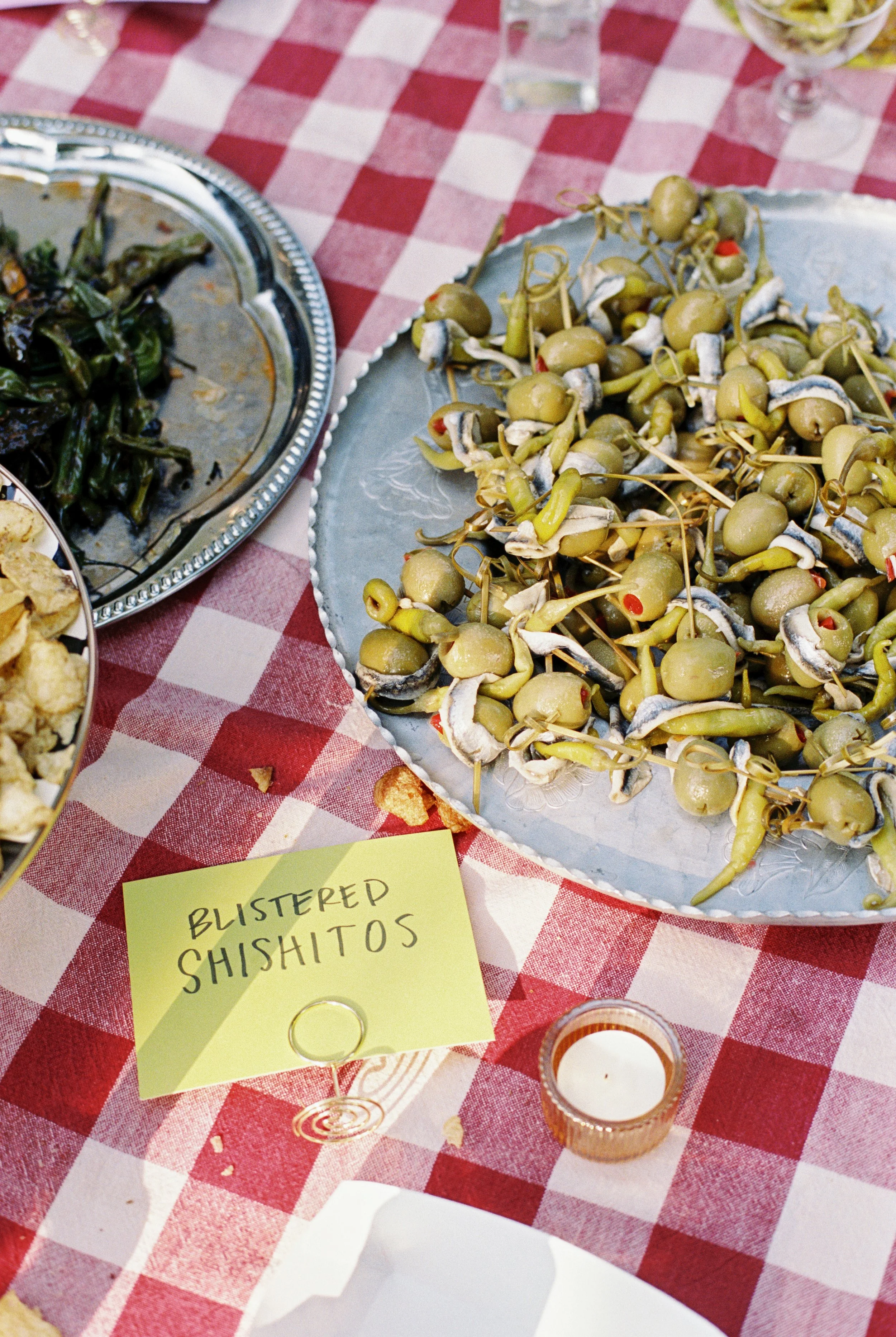 A plate of blistered shishitos peppers on a table with a red and white checkered tablecloth, next to a small candle and a yellow sign labeled 'Blistered Shishitos'. There are other dishes partially visible.