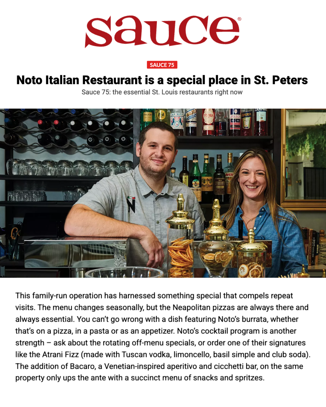 A photo of two smiling people, a man and a woman, behind a bar with bottles and jars of citrus slices and herbs, representing a lively restaurant or bar setting.