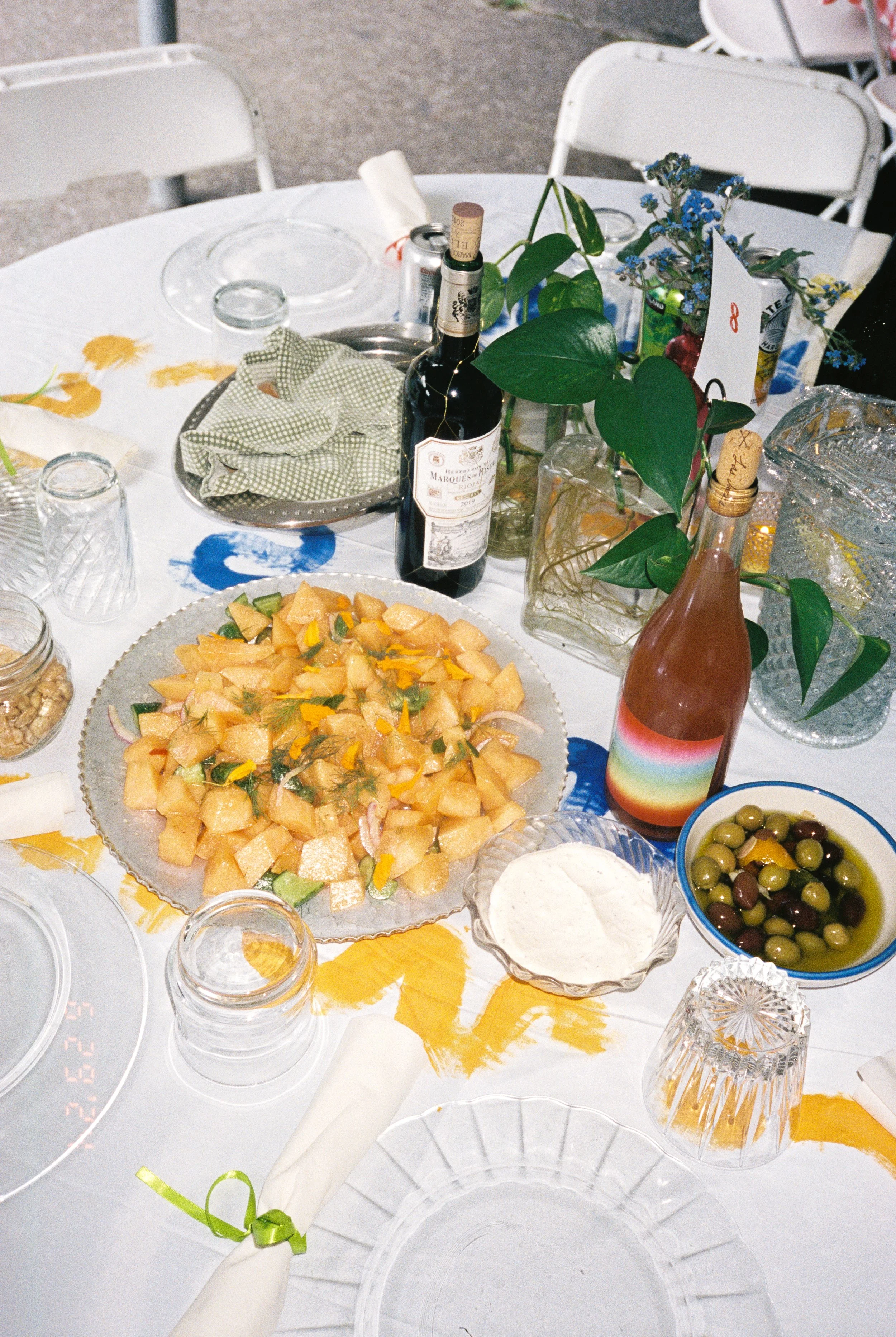 A round table set for a meal with various dishes, including a large plate of chopped fruit, a bowl of olives in oil, a bowl of creamy dip, and bottles of wine and rosé. The table has glasses, napkins, and decorative flowers.