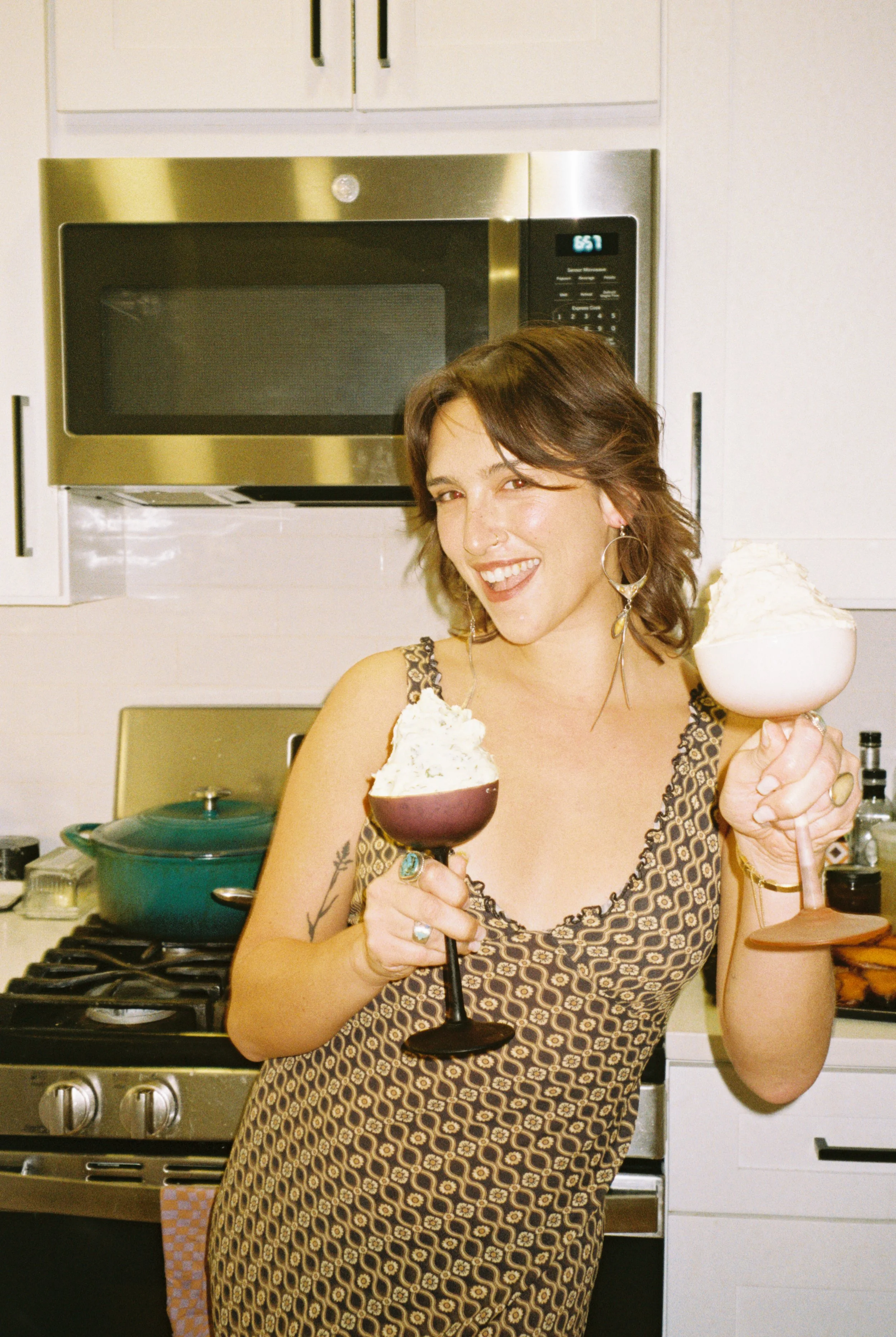 A woman in a patterned dress holding two bowls of whipped cream and chocolate mousse in a kitchen with white cabinets and a microwave.