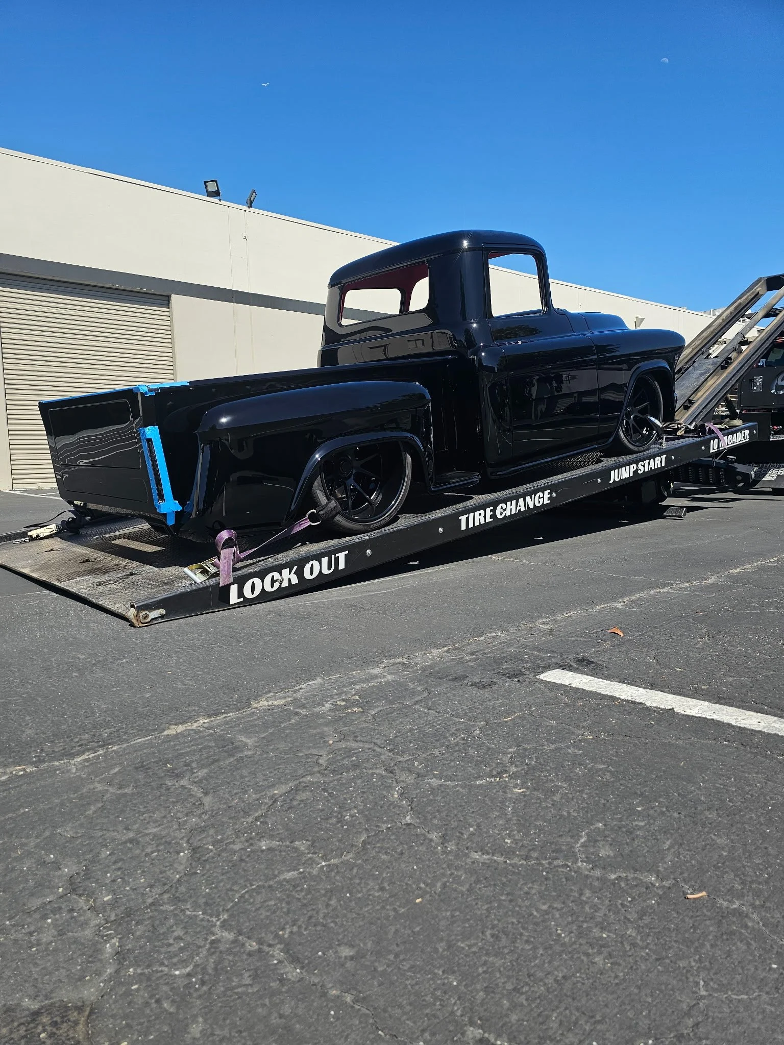 A black classic truck being transported on a flatbed truck. The flatbed has labels for lockout, tire change, jump start, and load guider.