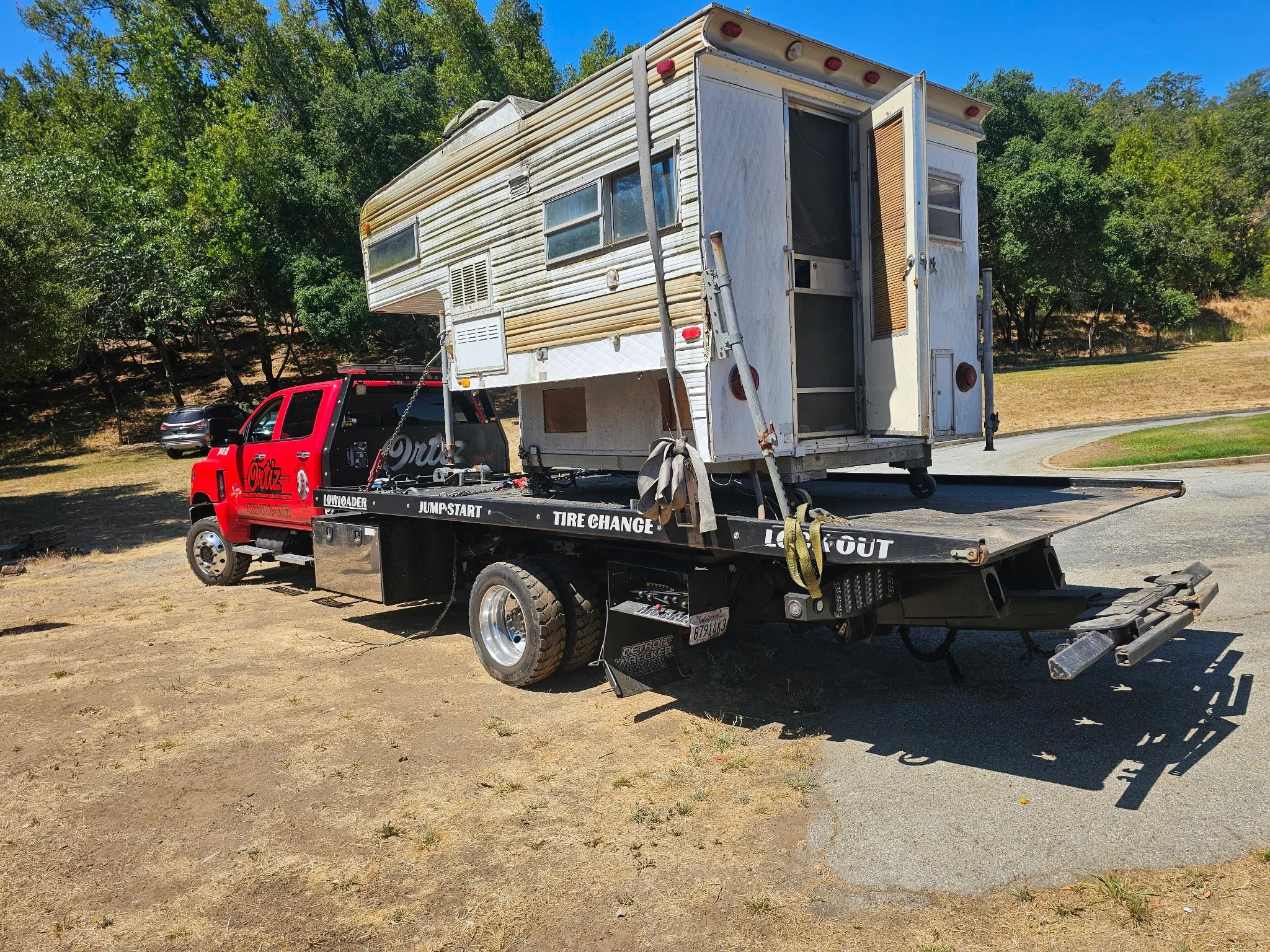 A red tow truck with a flatbed carrying an old, weathered camper trailer in a grassy area with trees in the background.