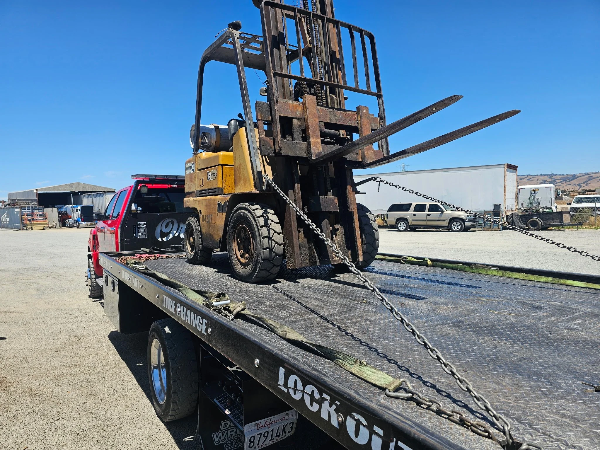 A forklift secured on the back of a flatbed truck in an industrial lot with trucks and warehouses in the background.