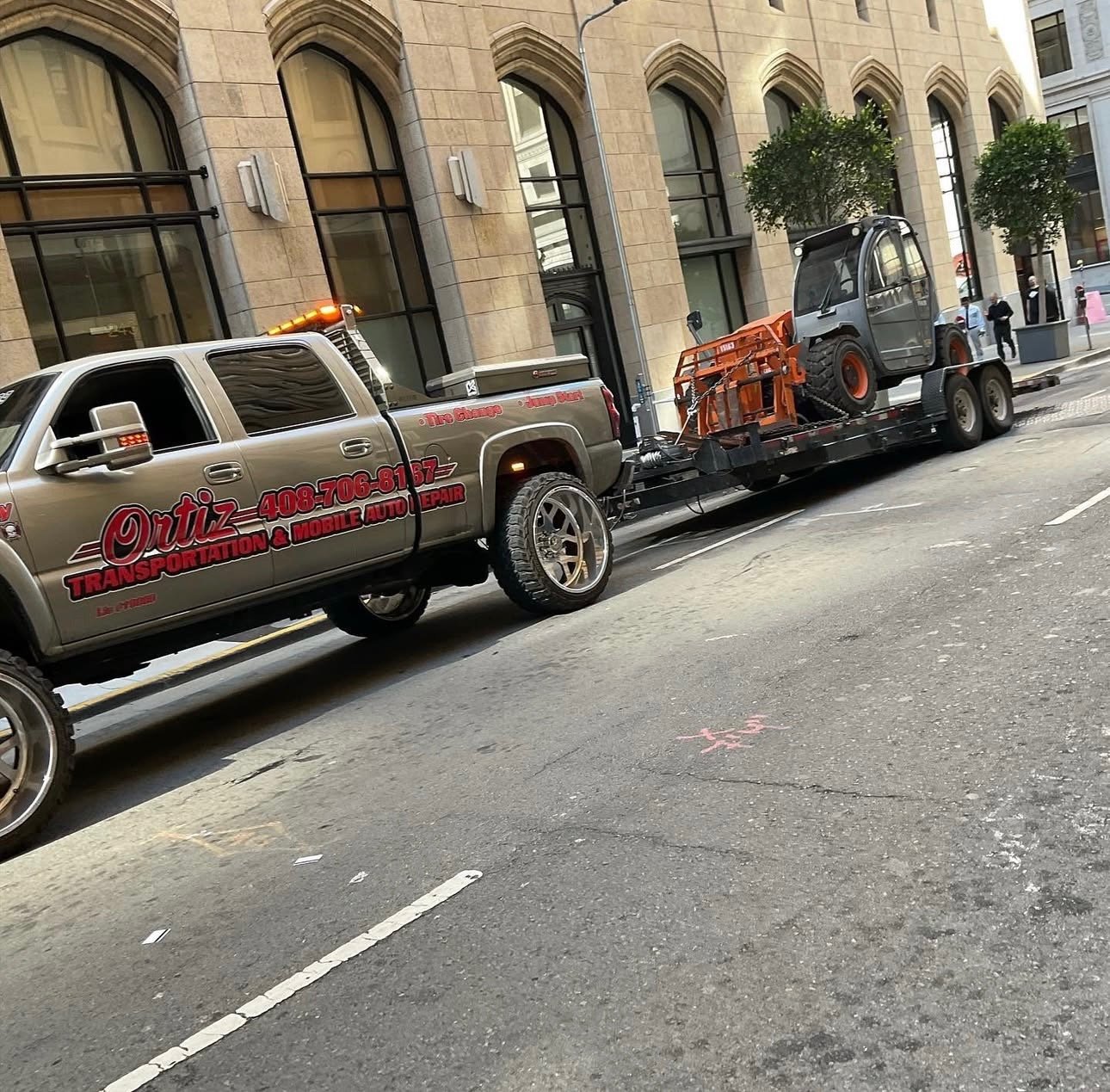 A tow truck transporting a small construction vehicle on a city street with brick building facades and trees in the background.