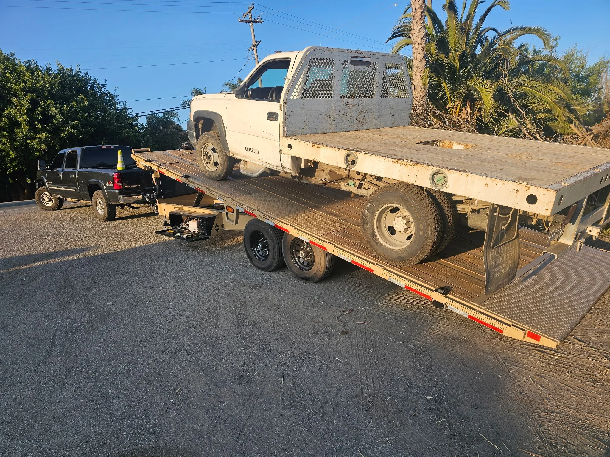 A flatbed tow truck carrying a white pickup truck with a metal mesh dividing panel in the cargo area. The tow truck is on a road with a black pickup truck behind it.