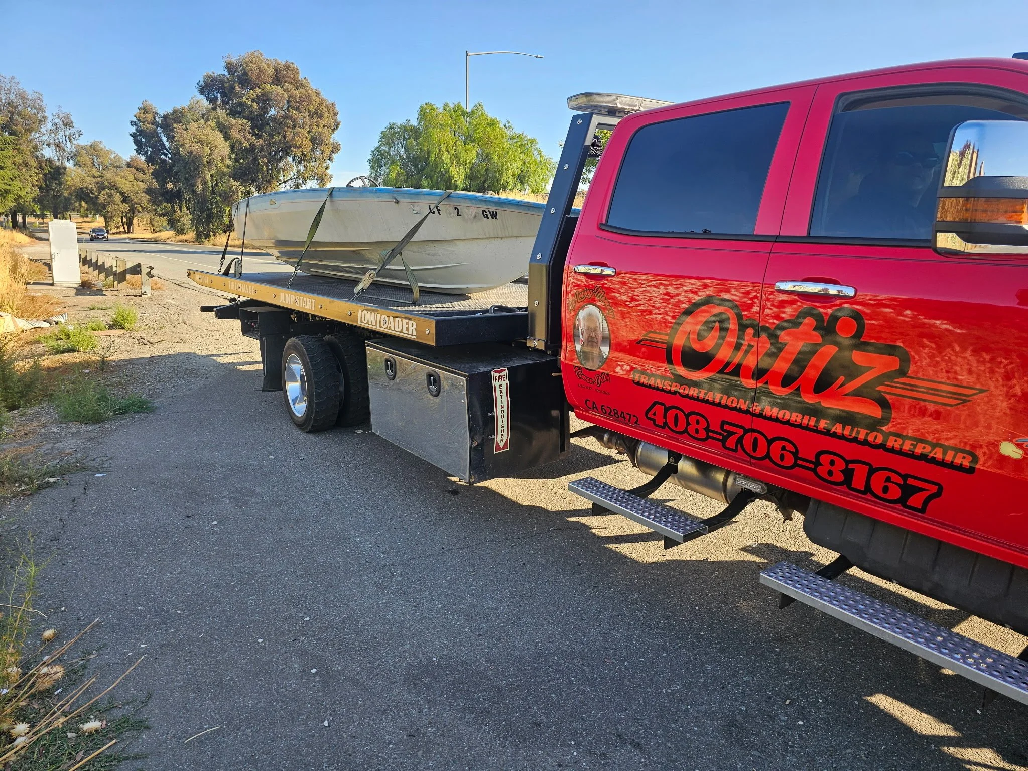 Red transportation service truck with custom logo and phone number, towing a boat on a trailer along a rural road during daytime.