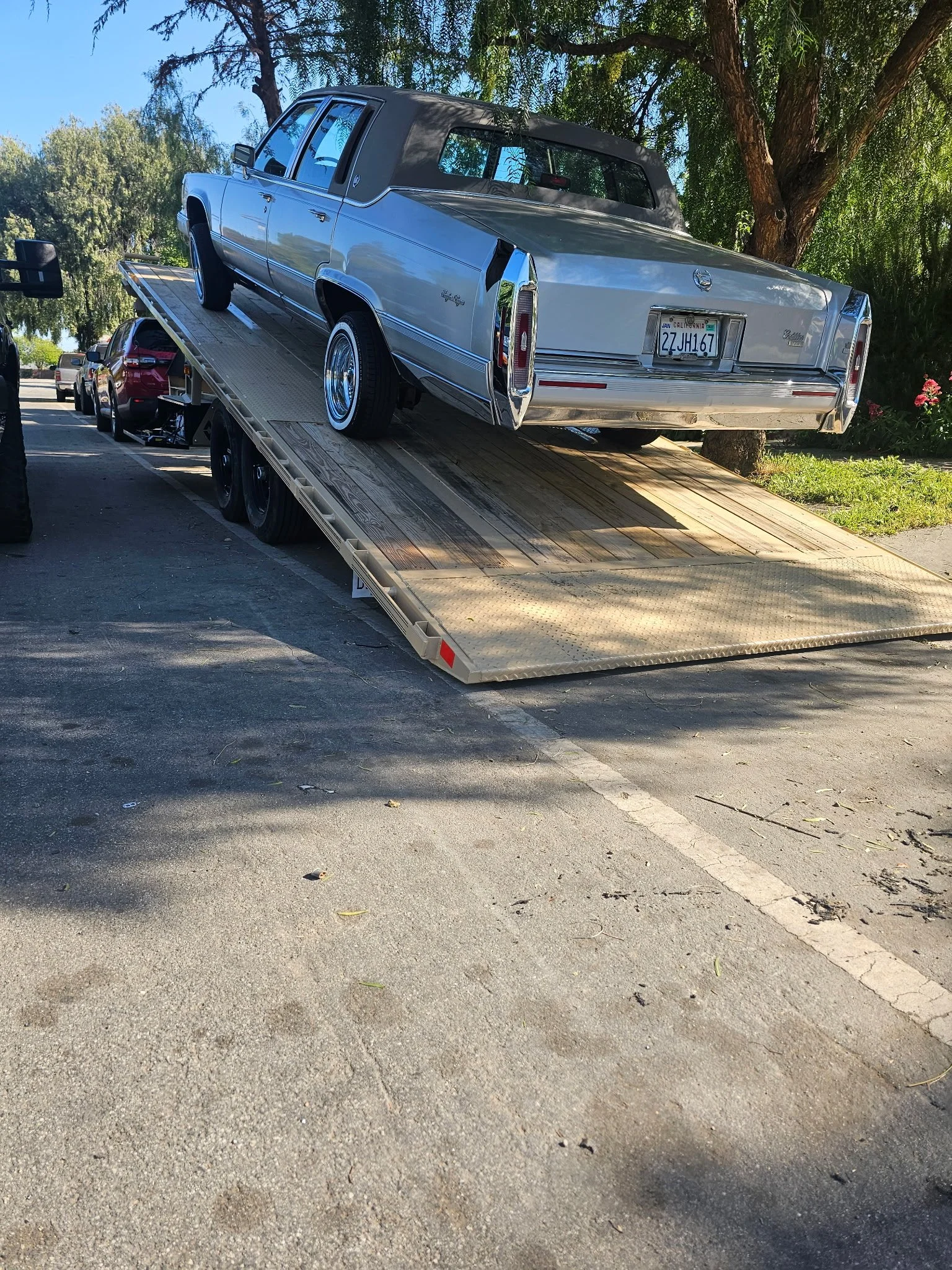 A silver classic car loaded on a flatbed tow truck in parking lot with trees in background