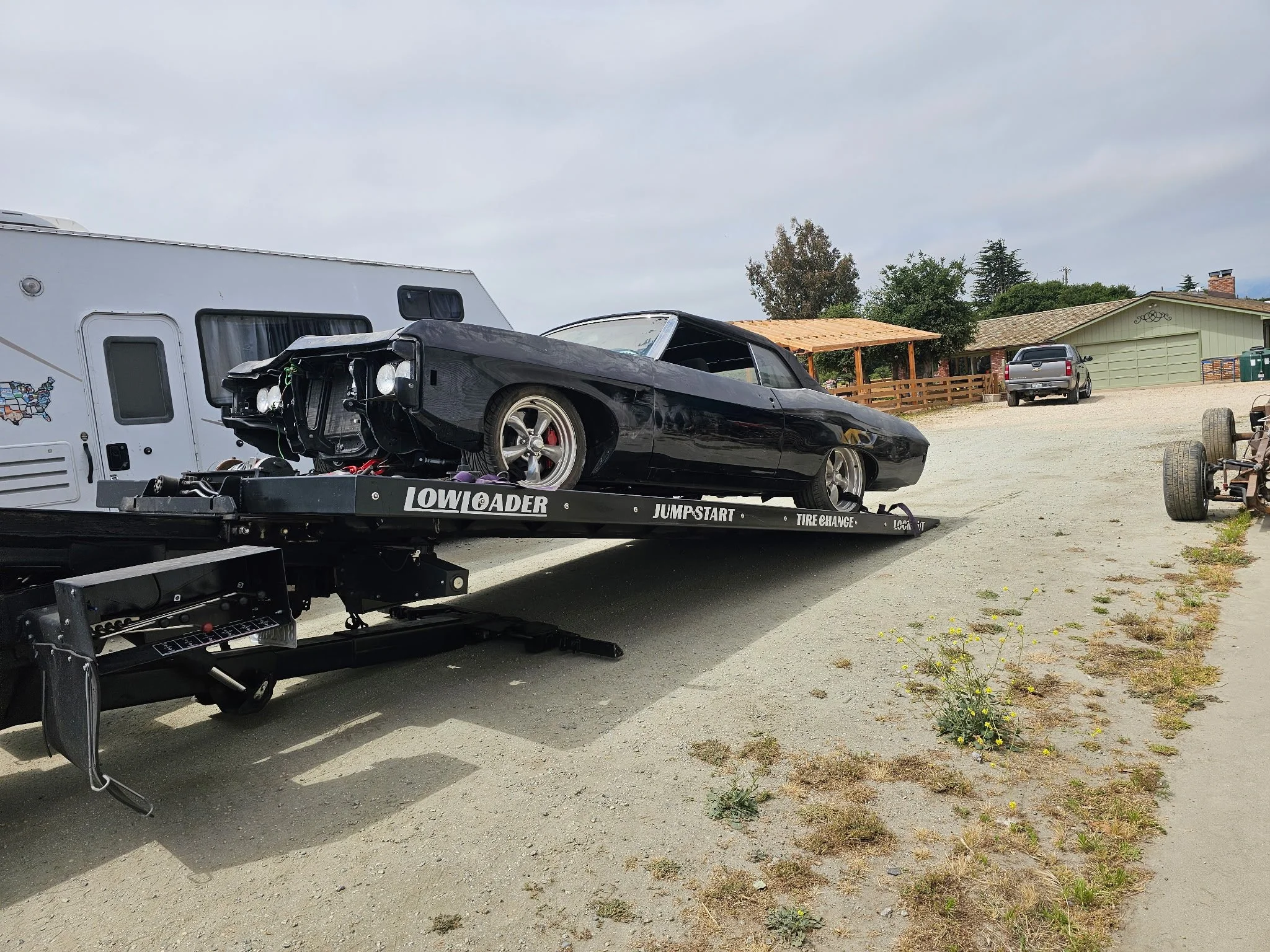 Black car on a trailer labeled 'LOW LOADER' parked on a dirt lot, with a white RV trailer and residential houses in the background.