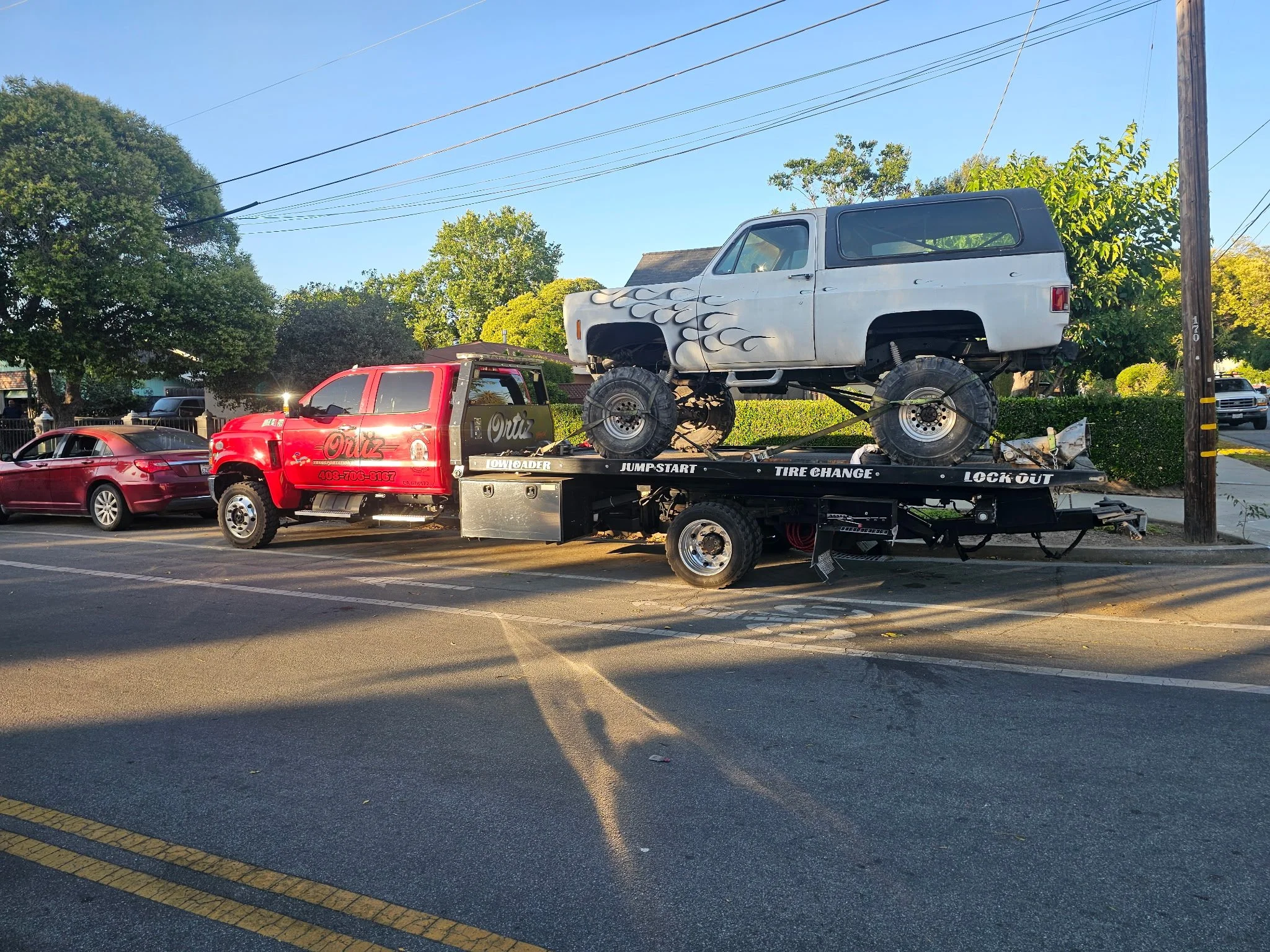 A red tow truck carrying a vintage off-road vehicle with large tires, painted in gray with flame decals, on its flatbed on a city street during daylight.