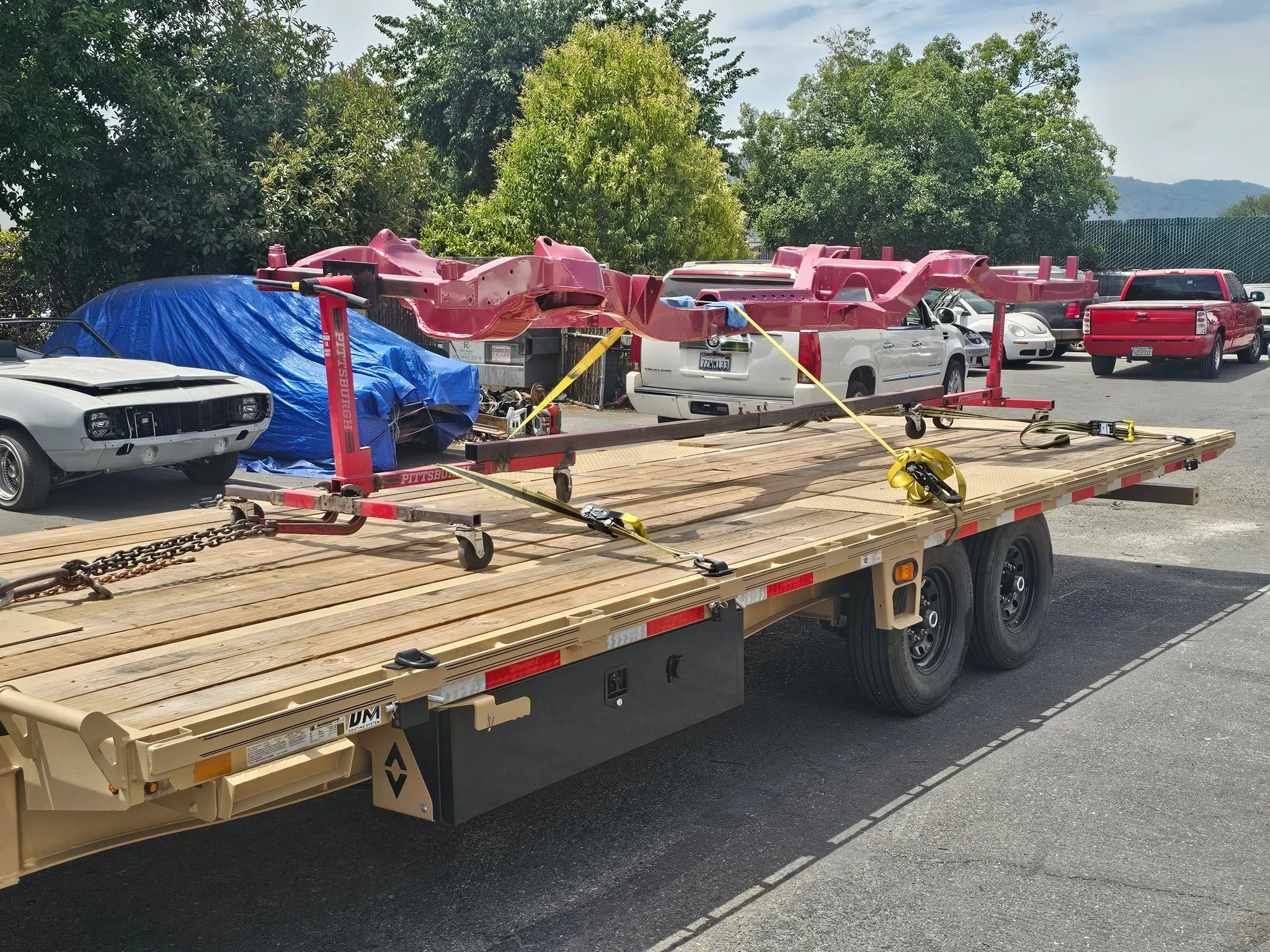 Flatbed truck with a pink car frame secured with straps, parked in a parking lot with various vehicles and green trees in the background.