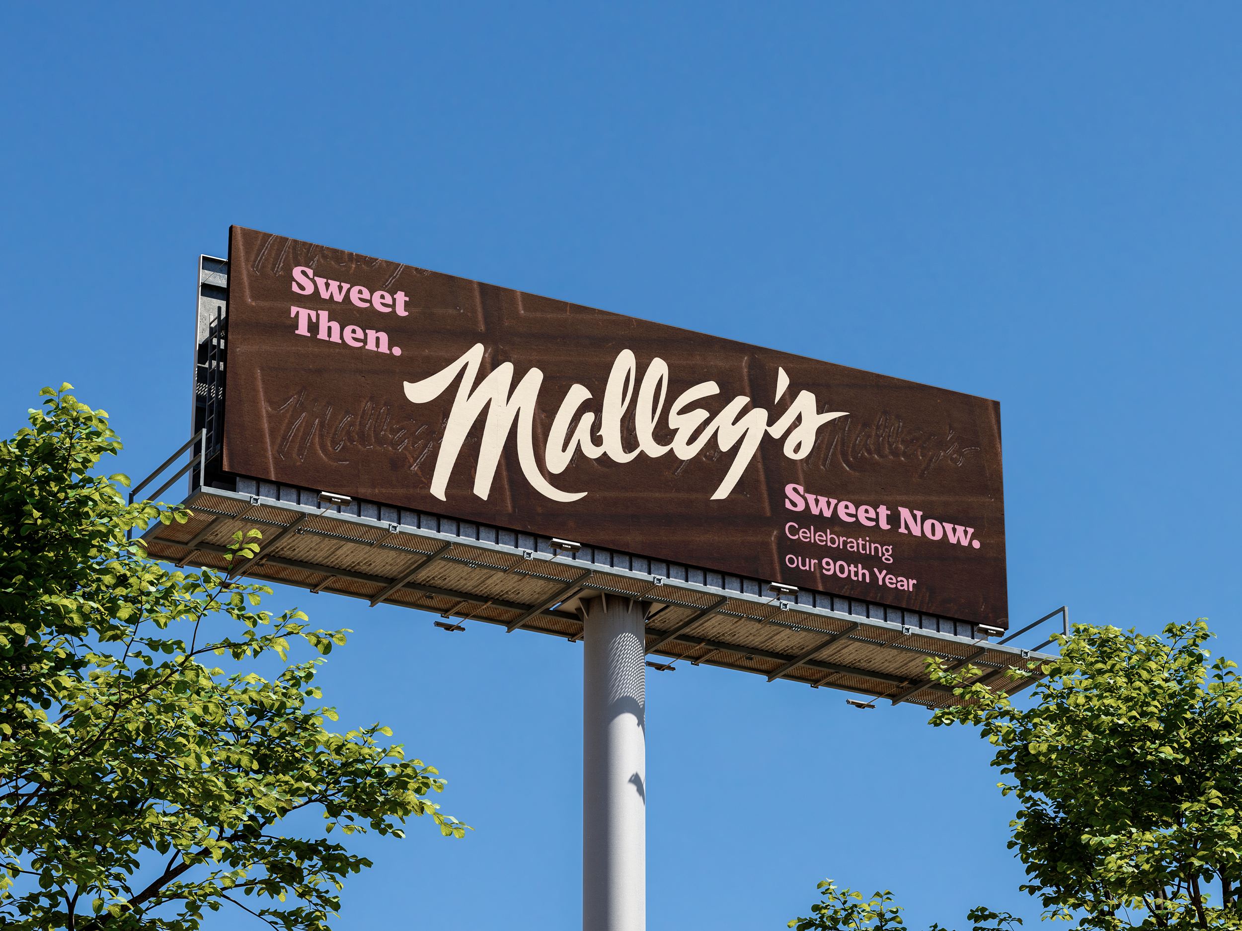 Billboard advertising Mallory's chocolates with the message 'Sweet Then. Sweet Now. Celebrating our 90th Year' against a blue sky with green trees in the foreground.