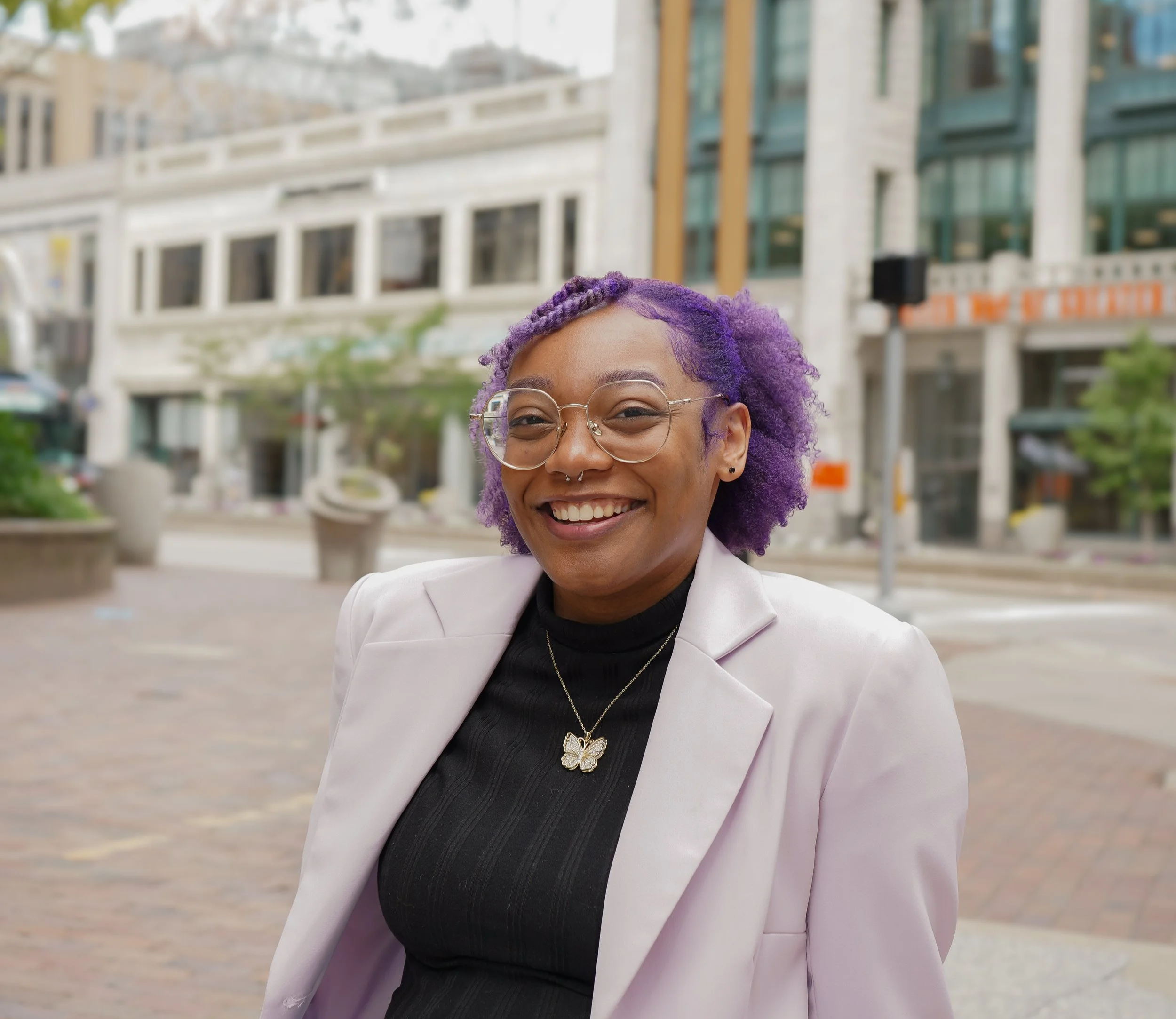 A woman with curly purple hair, glasses, a septum piercing, and a butterfly necklace, smiling outdoors in an urban setting wearing a black top and a light blazer.