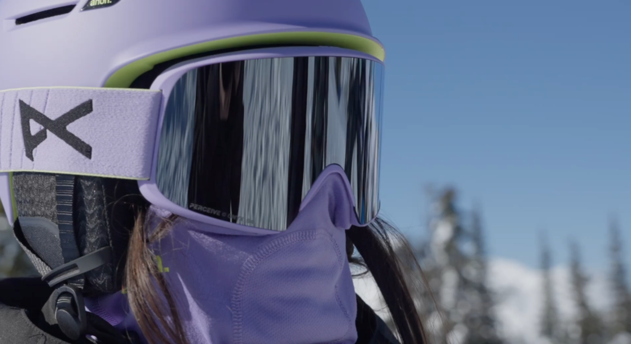 Person wearing ski goggles, purple helmet, and purple ski gear outside in a snowy landscape with snow-covered trees and a clear blue sky.