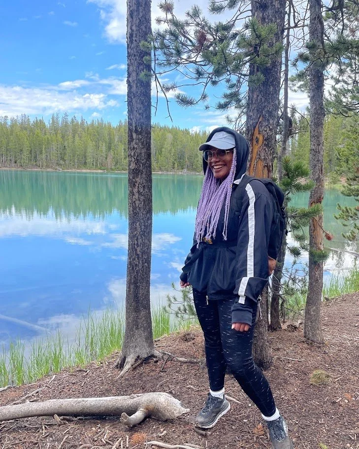A woman standing on a forest trail near a lake, smiling, wearing a hoodie, cap, glasses, and backpack, with trees and water in the background.