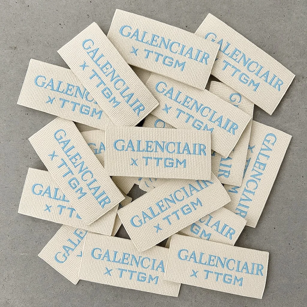 Multiple white fabric tags with blue text reading 'GALENCIAIR x TTGM' on a gray surface.