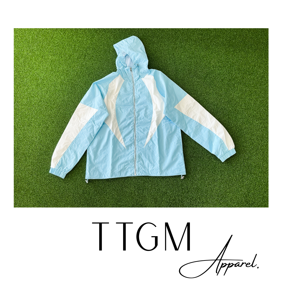 Light blue and white color-block windbreaker jacket with a hood, laid flat on green grass background, with text "TTGM Apparel" at the bottom.