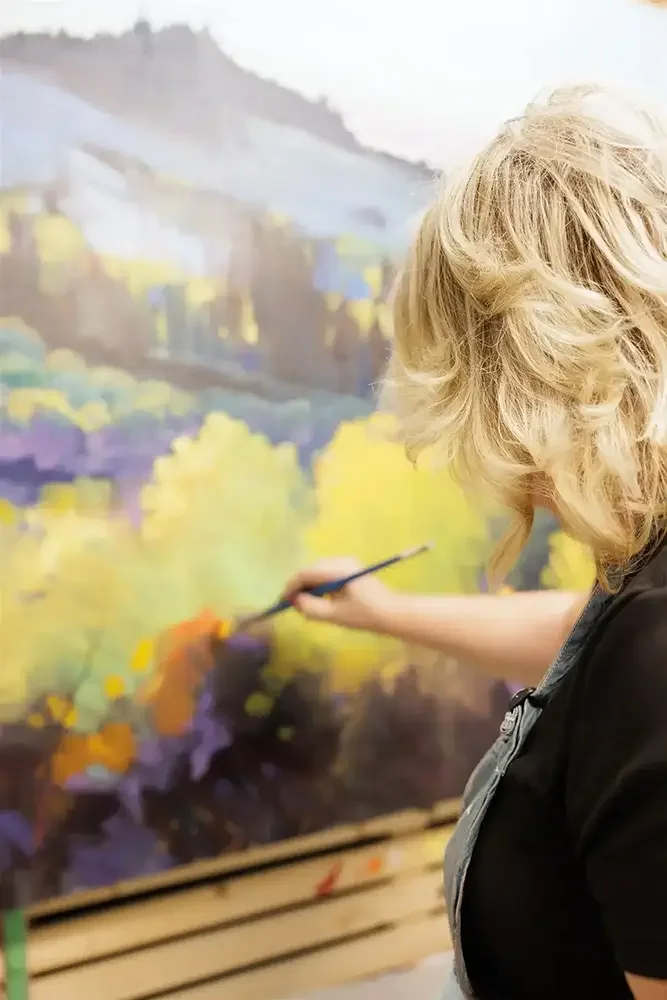 An artist with curly blonde hair painting a colorful landscape on a large canvas in an art studio.
