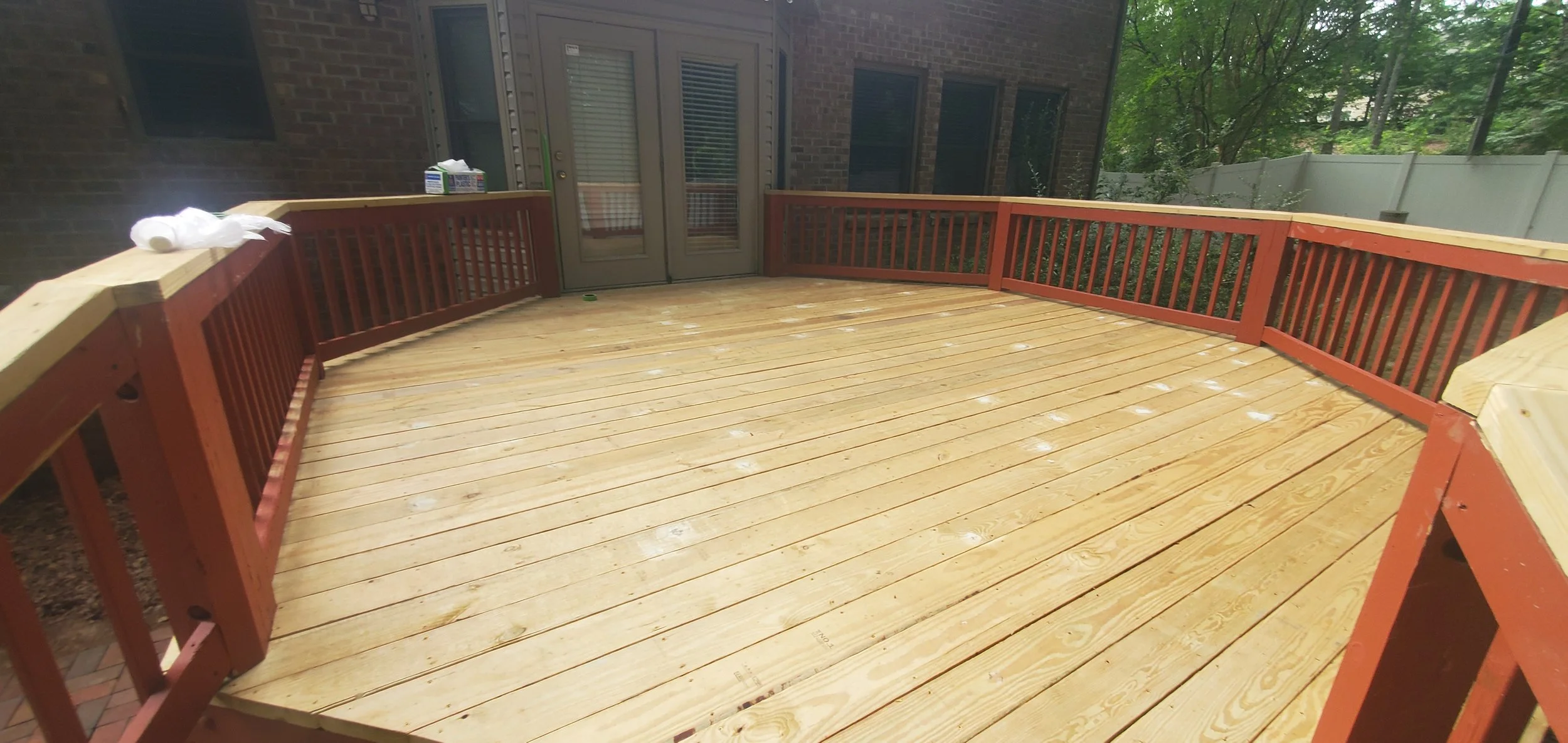Newly constructed wooden deck with railing attached to the back of a brick house, with tools and materials visible on top of the railing.