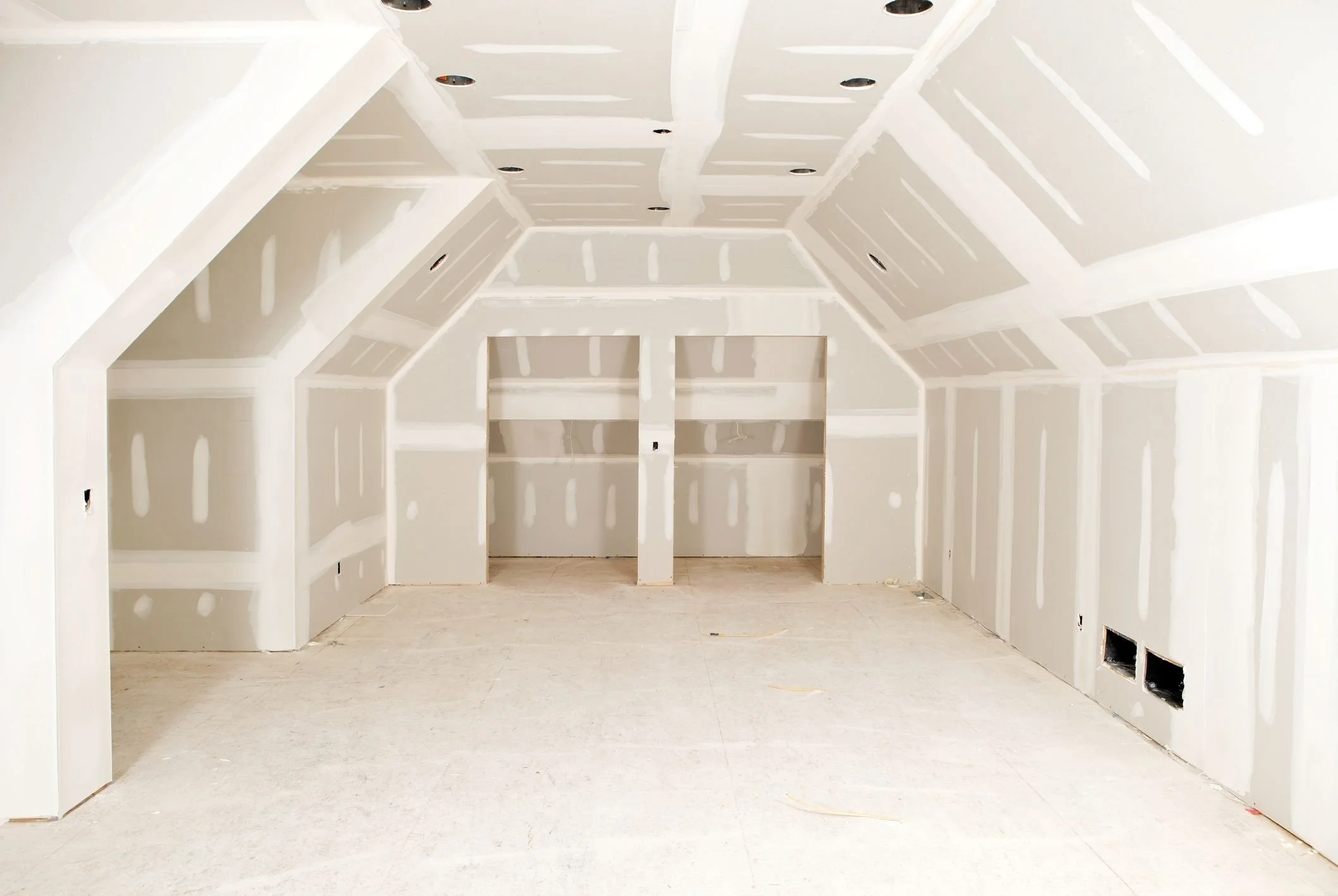 An unfinished room with drywall installed, showing taped seams and no flooring, ceiling lights installed, and two small openings in the lower right wall.