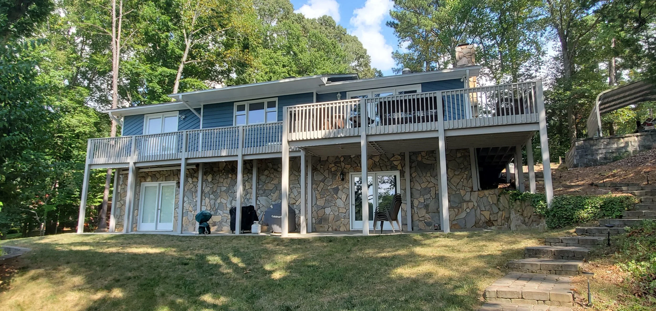A two-story house with a stone foundation, blue siding, a large wooden deck on the second floor, and glass sliding doors on the lower level set in a wooded area.